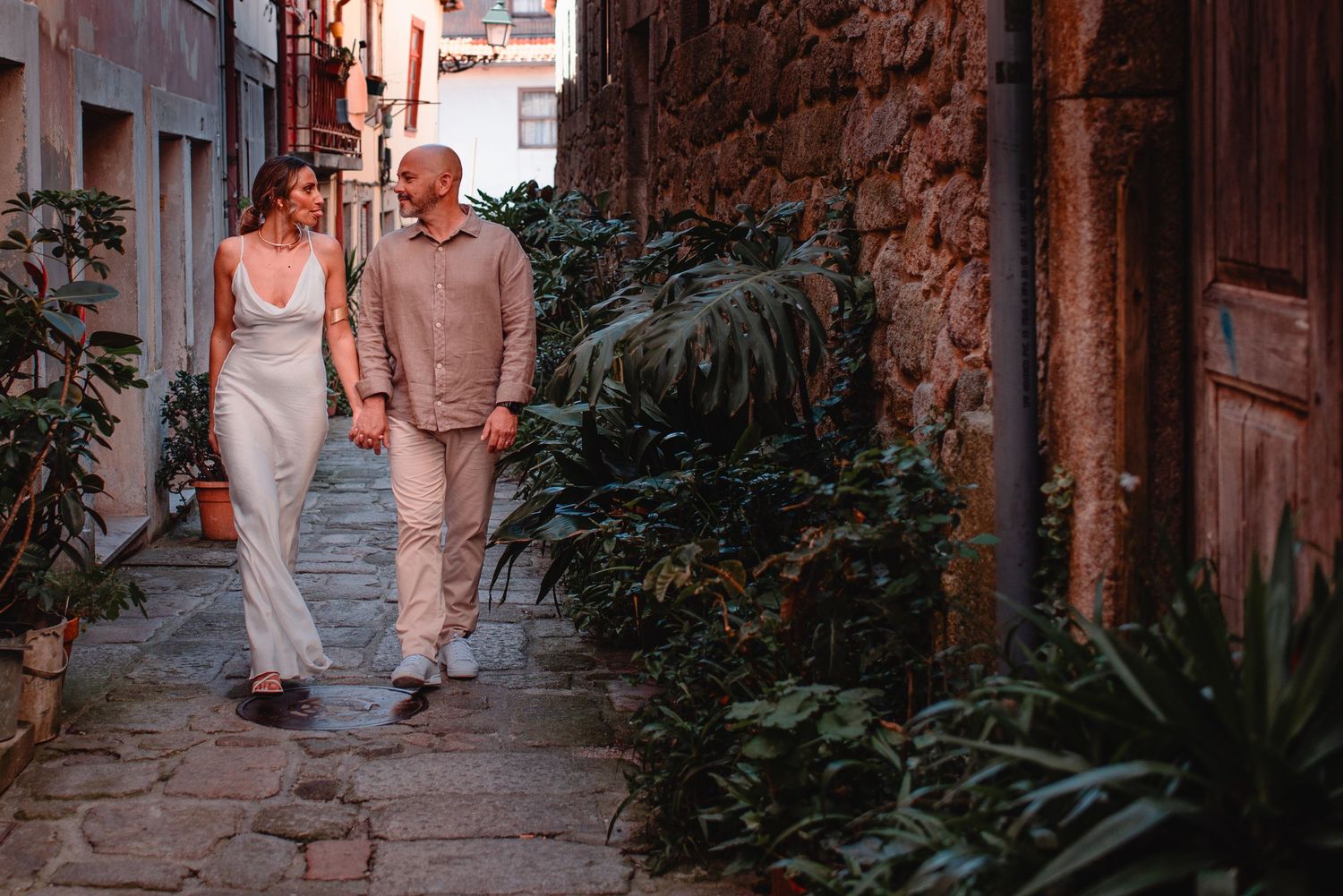 Two people share a tender moment in a charming Mediterranean alley with potted plants.
