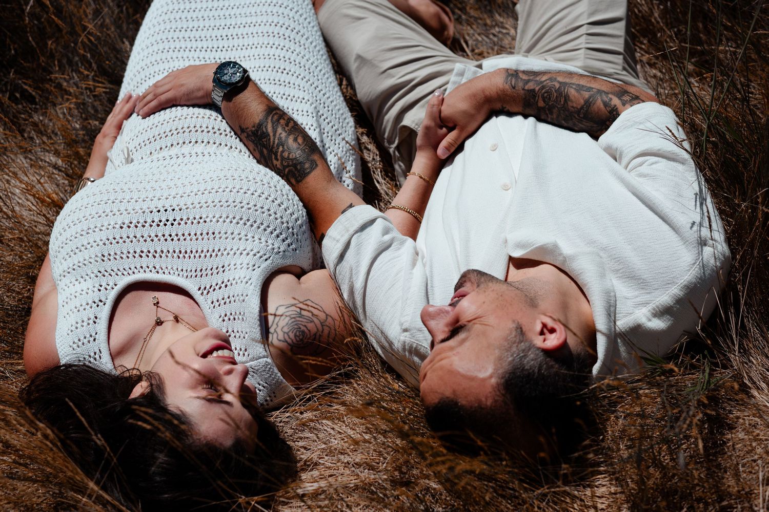 Couple lying in tall grass wearing white outfits and laughing together.