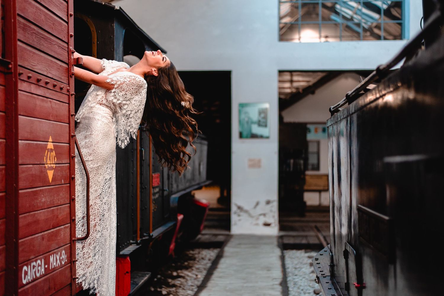 Person in white lace wedding dress posing dramatically against red vintage train car.