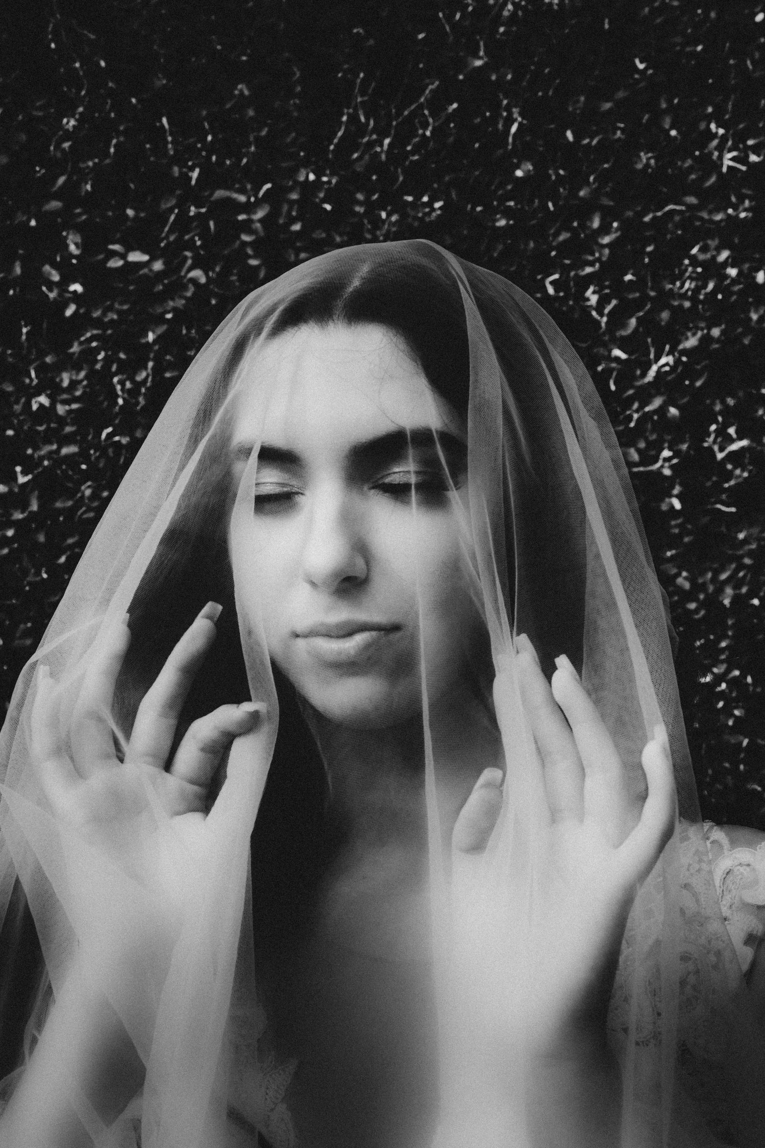 Black and white portrait of a bride with closed eyes, hands raised, covered by a sheer veil against dark foliage.