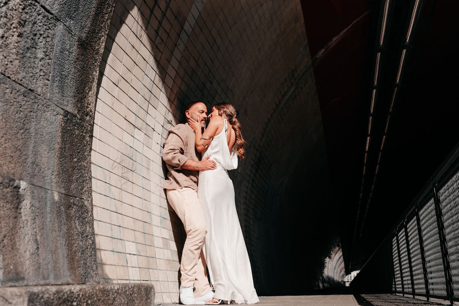 Bride and groom share a romantic moment under a stone archway tunnel in soft natural light.