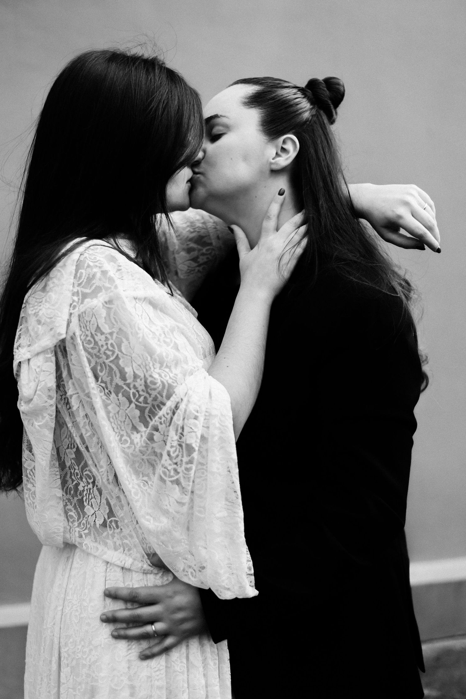 Two women sharing a kiss in black and white photo, one in white lace dress, other in black outfit.