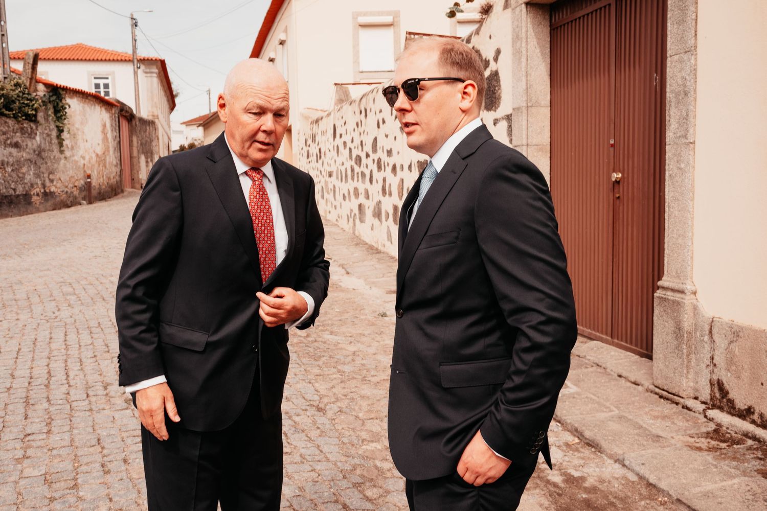 Two men in dark suits walking and talking on a cobblestone street in a European town.
