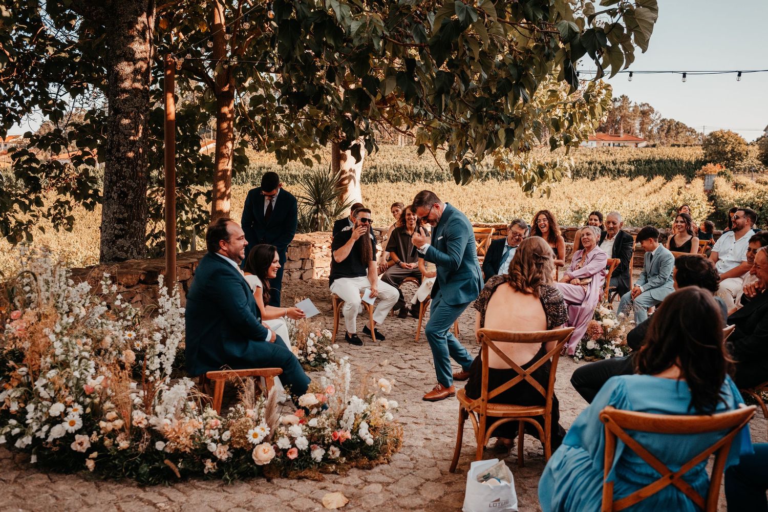 Outdoor wedding ceremony at a vineyard with guests seated among floral arrangements under string lights.
