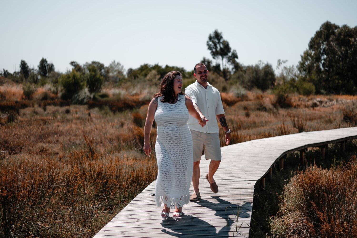 Couple walks hand in hand along wooden pathway near rustic white building.