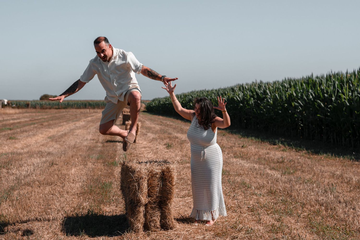 Romantic couple shares playful moment jumping over hay bale in field at sunset near cornfield.