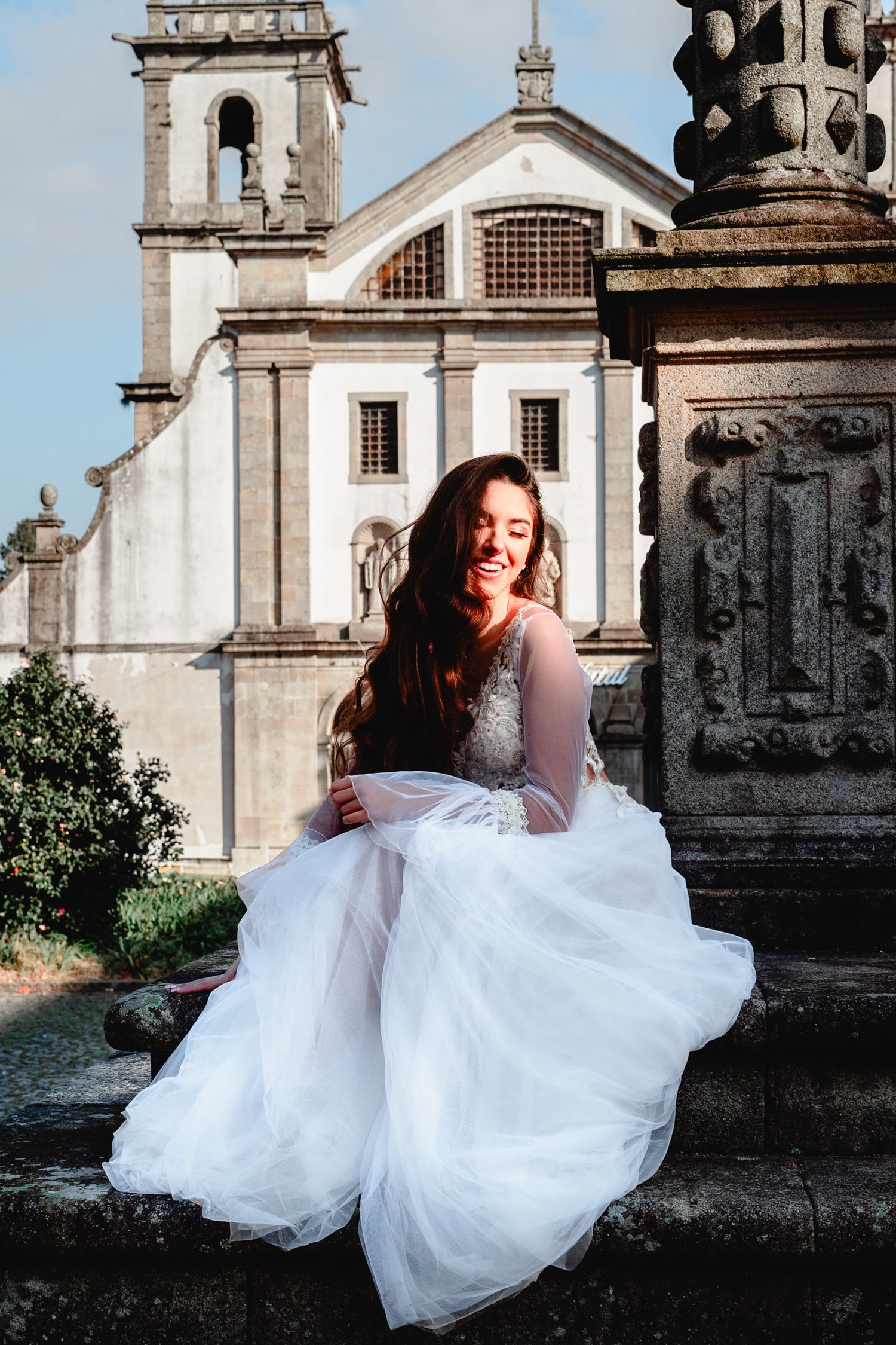 Bride in white lace gown sits gracefully on stone steps with classic church facade in background.