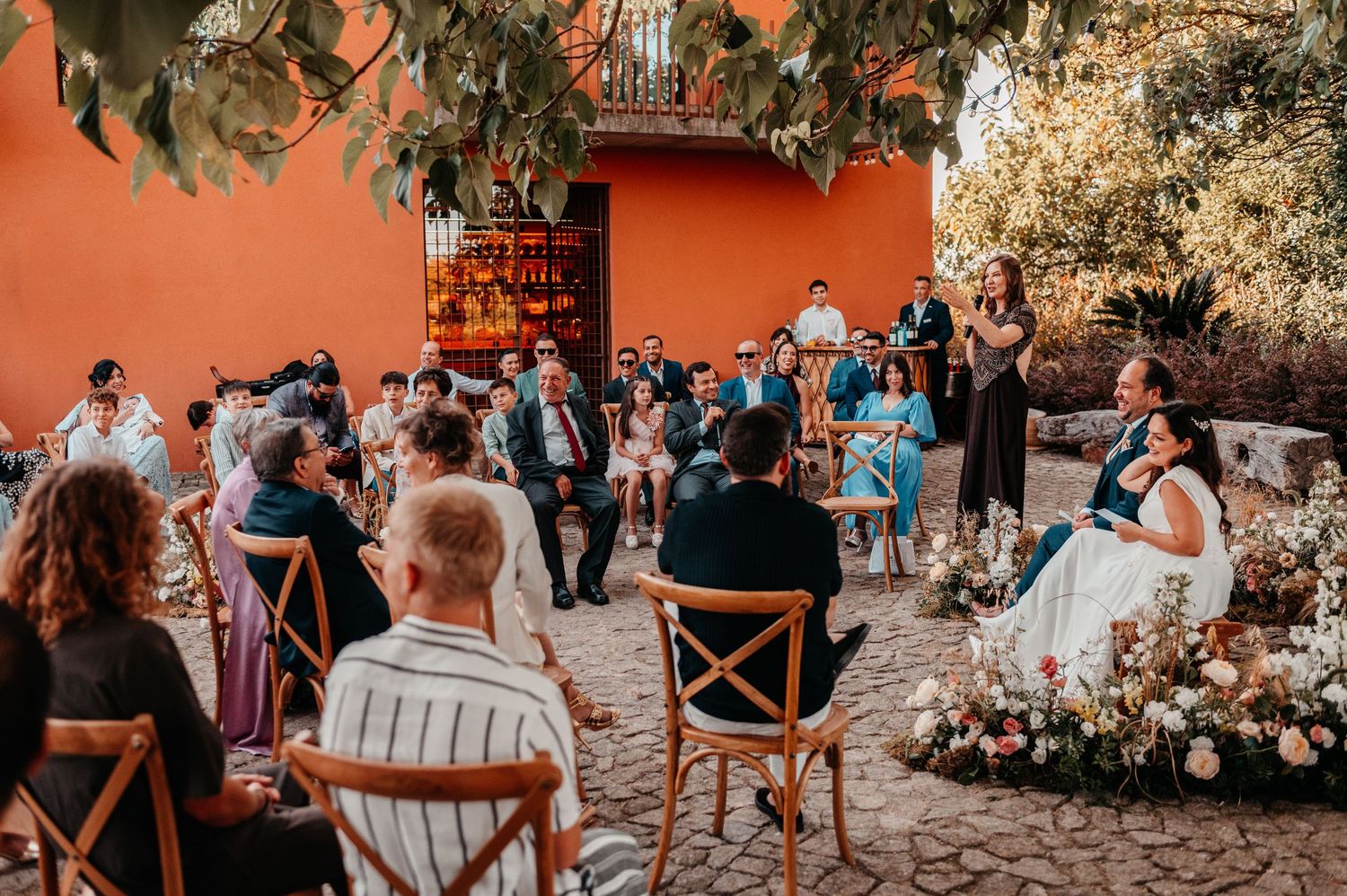 Outdoor wedding ceremony in a colorful courtyard with guests seated around the couple, surrounded by lush greenery and floral arrangements.