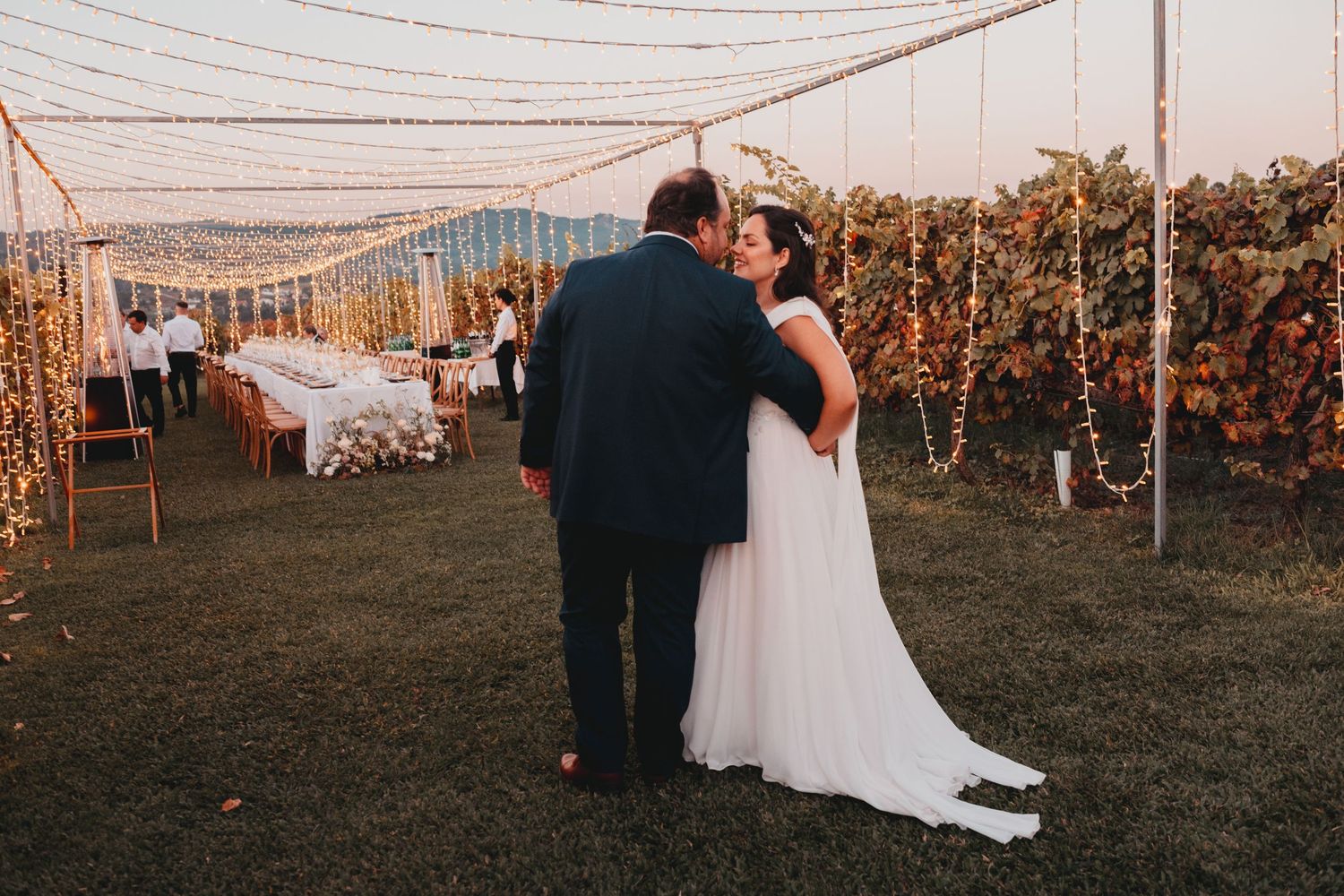 Bride and groom sharing a kiss at an outdoor vineyard wedding reception with string lights and mountain views.