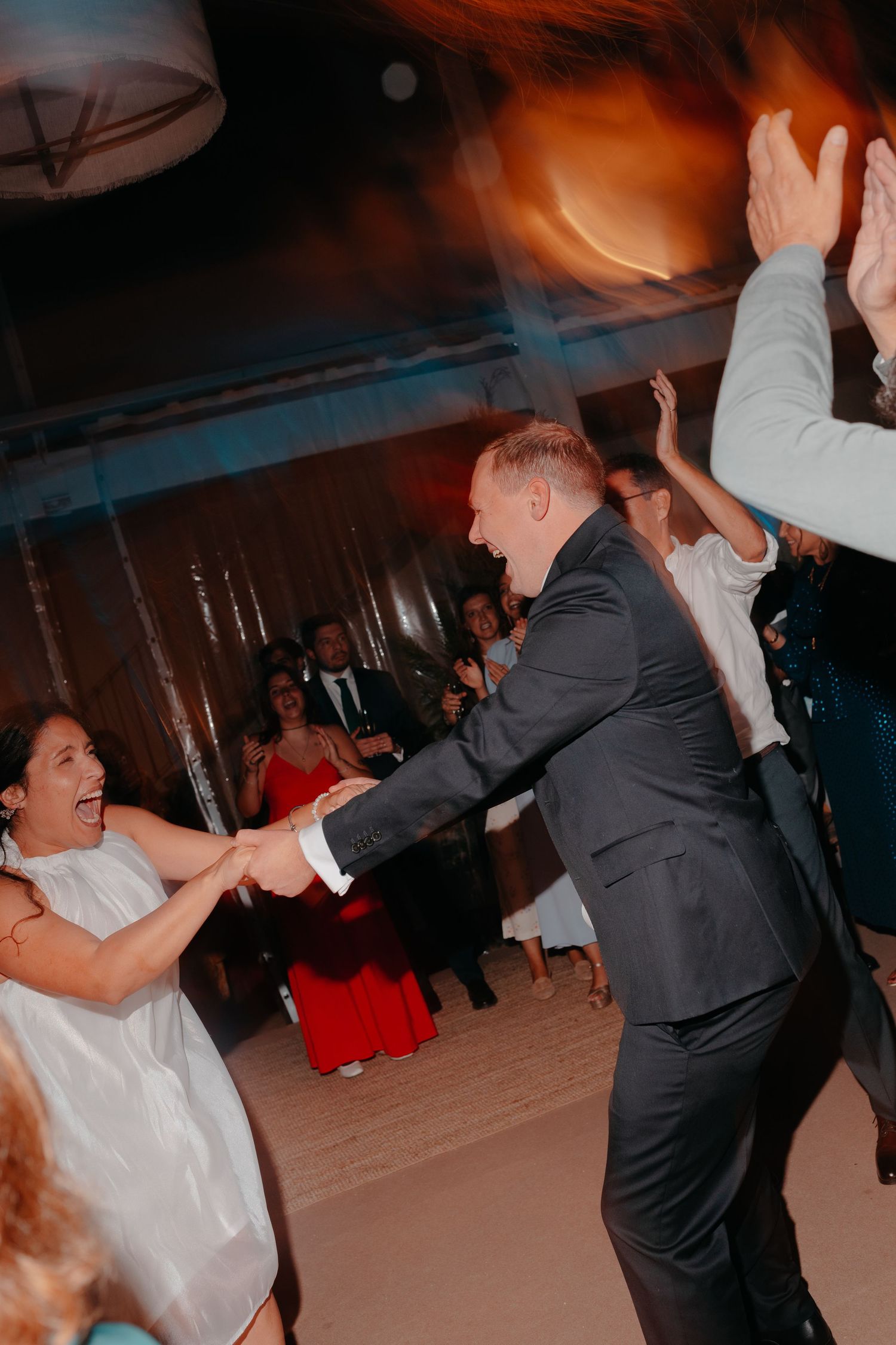 Joyful couple dancing energetically at a wedding reception, surrounded by guests in formal attire on a dimly lit dance floor.