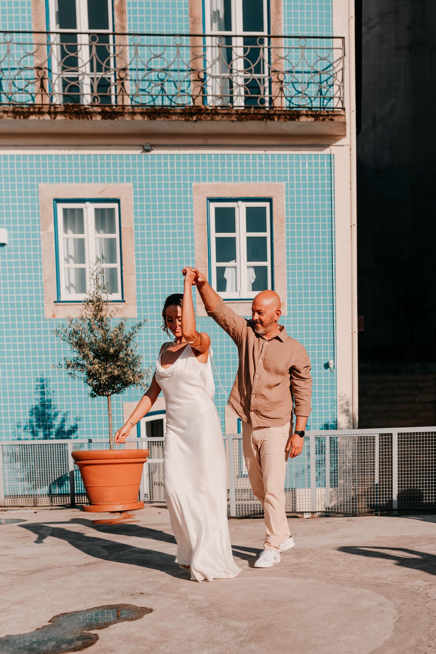 Couple dancing outdoors in front of a blue tiled building, woman in white dress, man in beige outfit.