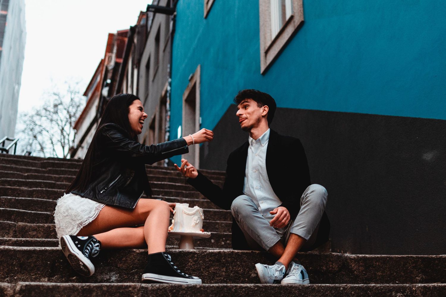 Young couple sitting on outdoor steps, laughing and talking near colorful buildings.