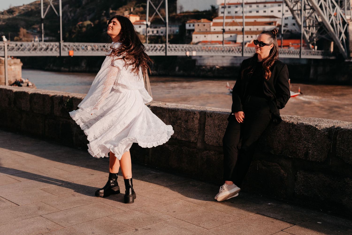 Couple posing by a riverside in Porto, woman in white dress twirling, man in black jacket seated on stone wall.