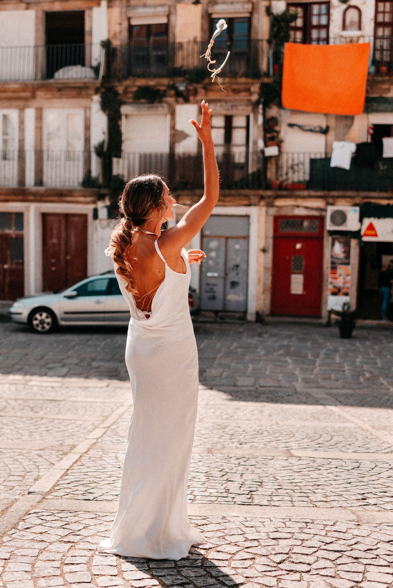 Bride in white backless gown tossing bouquet on cobblestone square in European city.