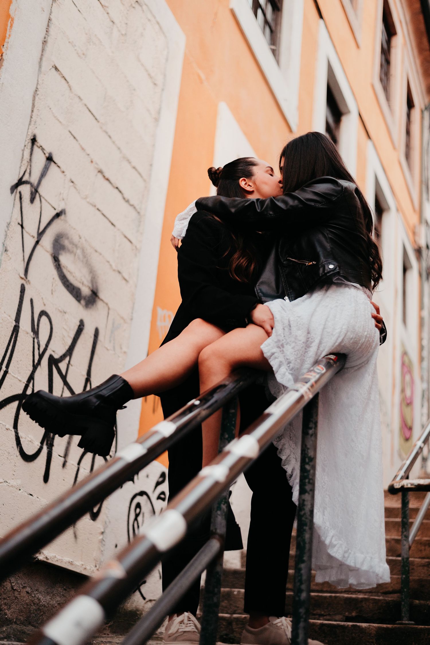 Two women sharing a romantic kiss on urban stairs with graffiti wall backdrop, one in white dress and leather jacket.