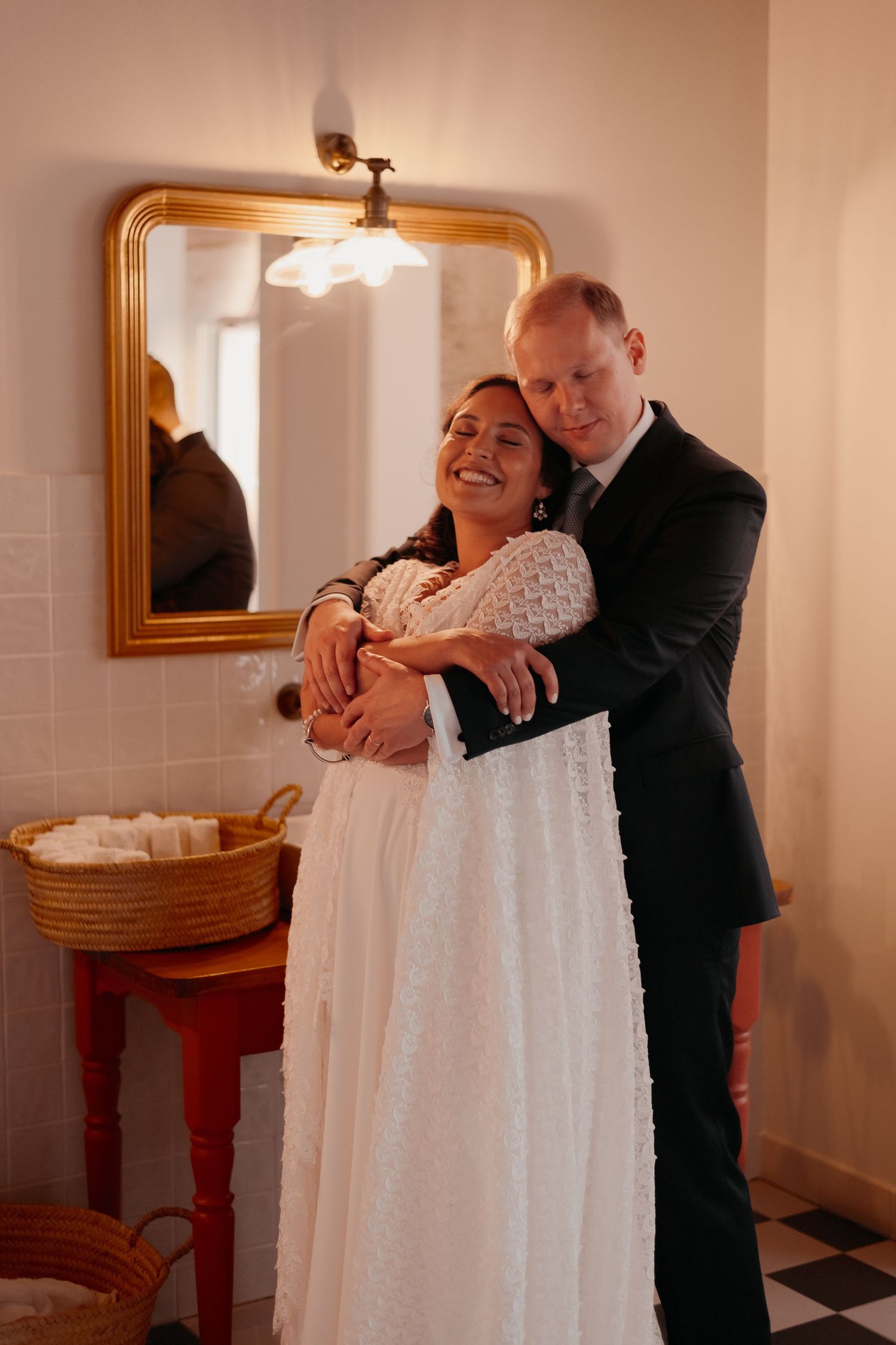 Bride in white dress embraced by groom in black suit, smiling in elegant room with gold mirror.