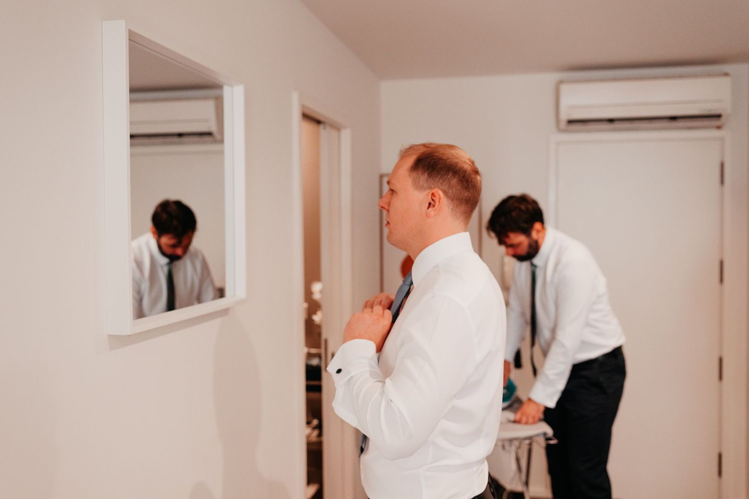 Groom adjusting his tie in a mirror reflection while a groomsman assists in a bright white room before a wedding.