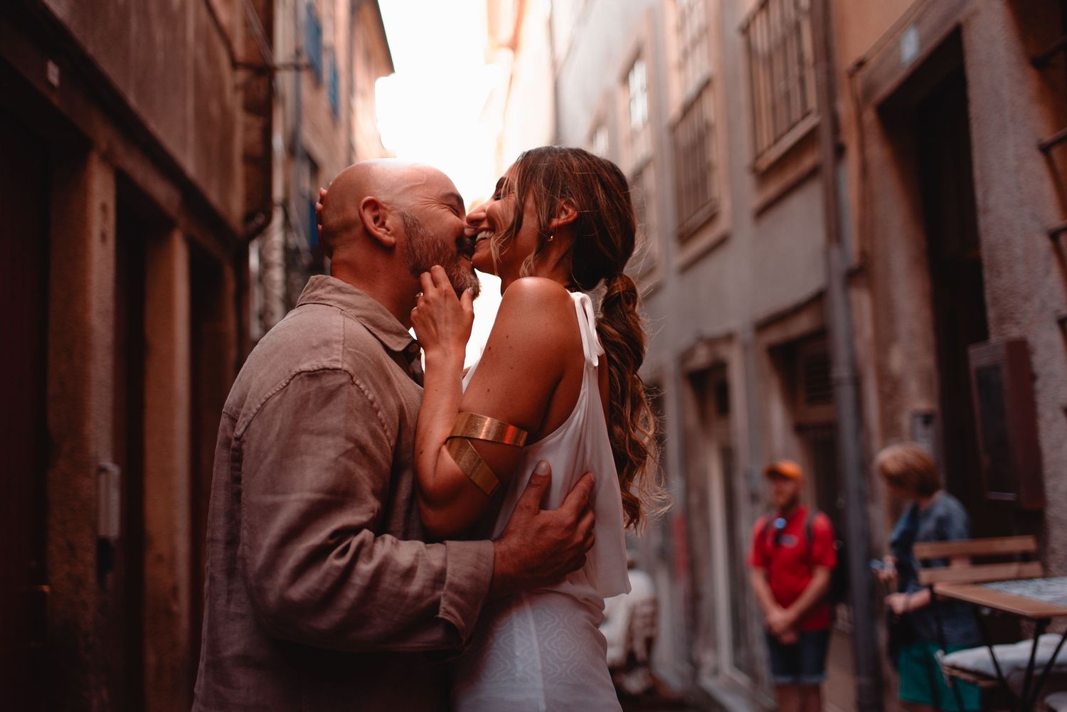 Couple sharing a romantic kiss in a narrow European alleyway, woman in white dress with gold bracelet.