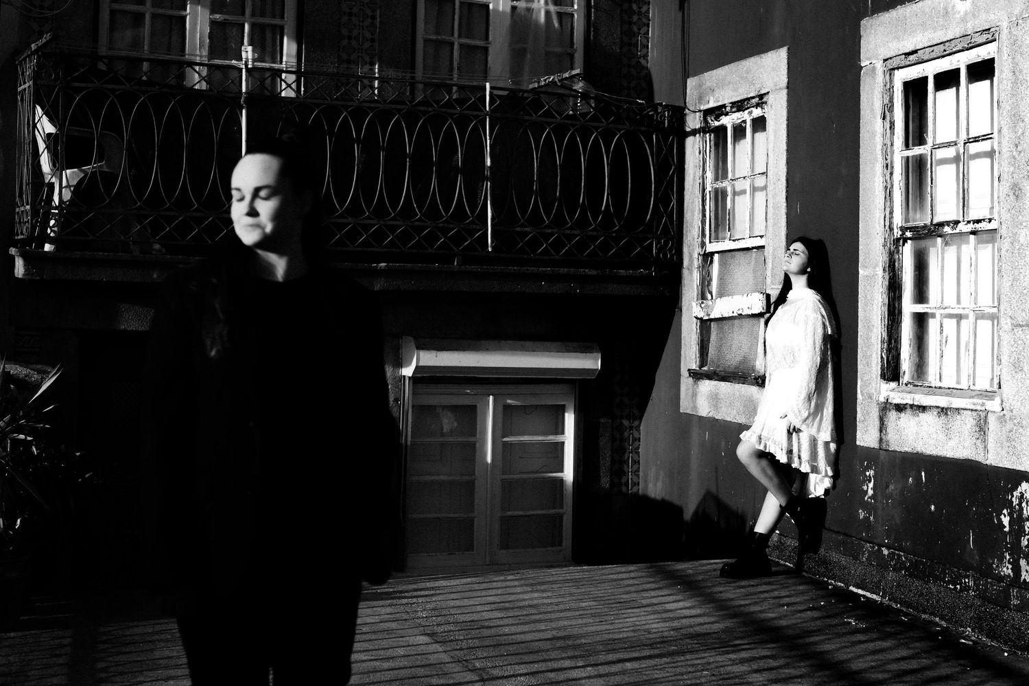 Black and white photo of two people outside a building with a balcony, one standing in shadow, one leaning against wall.