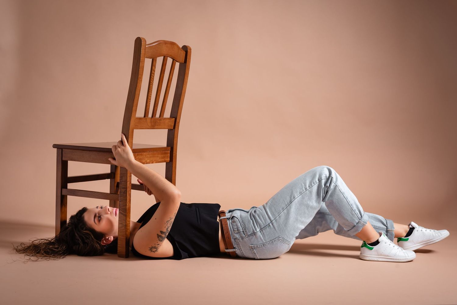 Person in casual attire lying on floor while balancing wooden chair above.