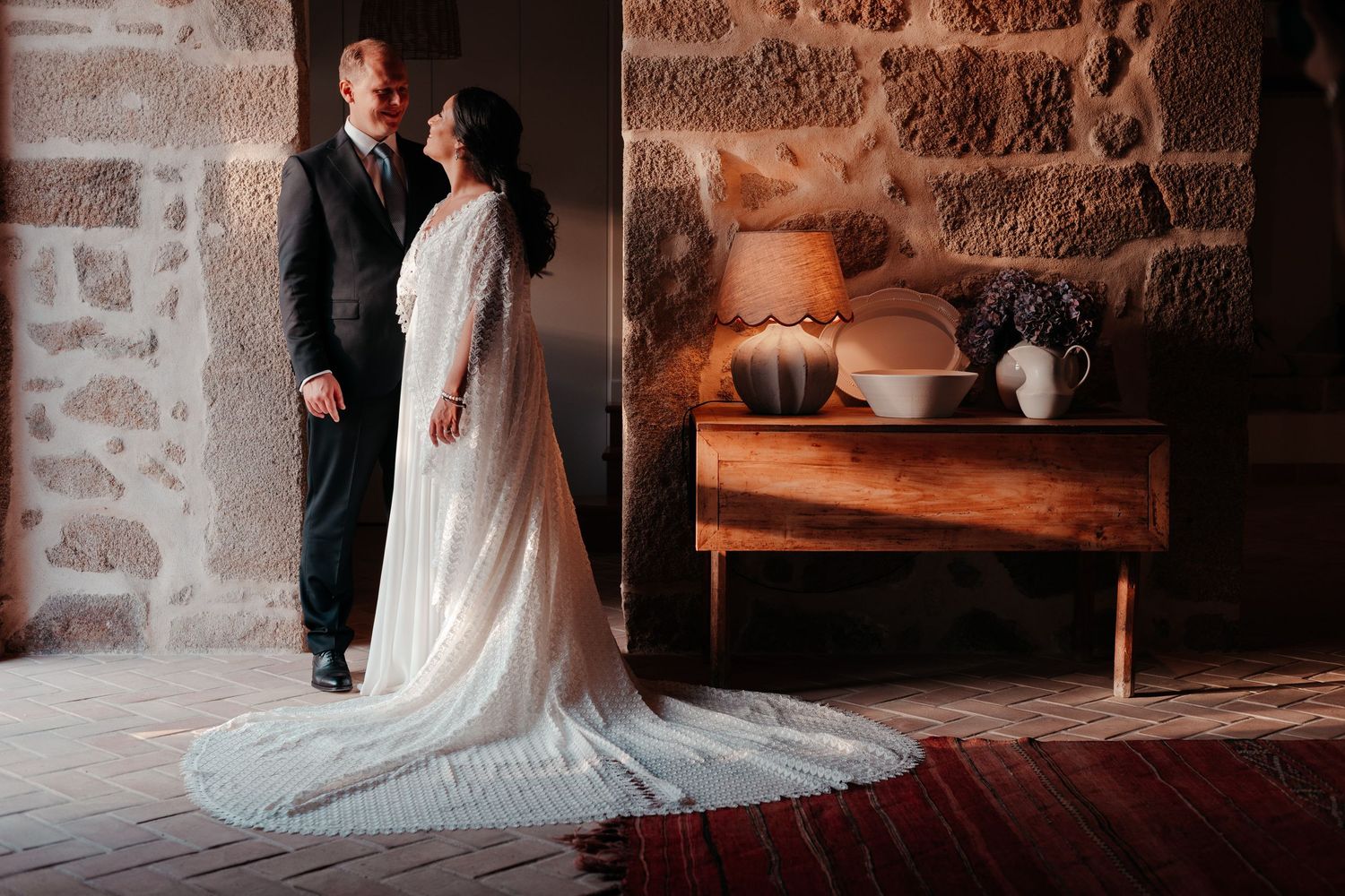 Bride in flowing white gown with train stands with groom in dark suit in rustic stone interior near wooden sideboard.