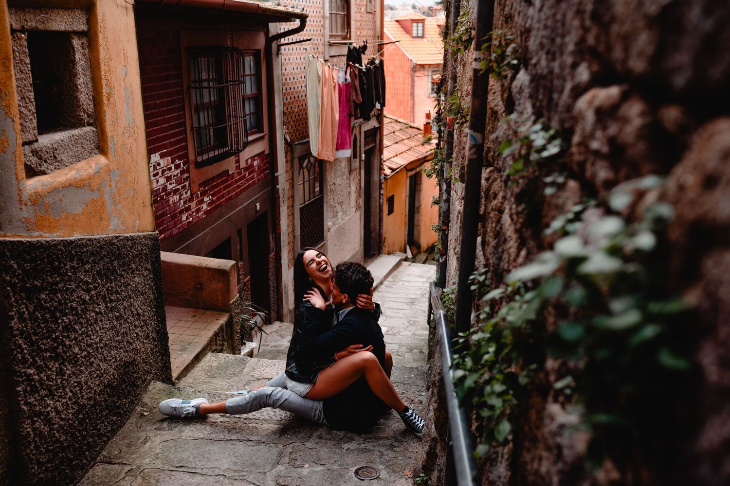 Two dancers rest on cobblestone steps in a narrow European alley, surrounded by rustic stone walls and colorful buildings.