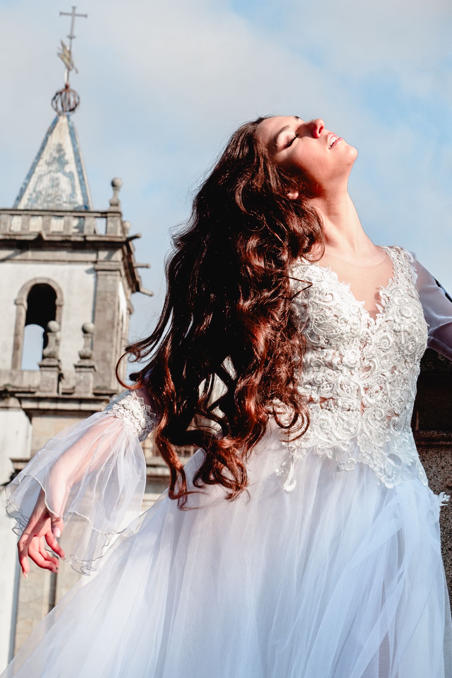 Artistic bridal portrait with church tower in background showing flowing hair and detailed lace dress.