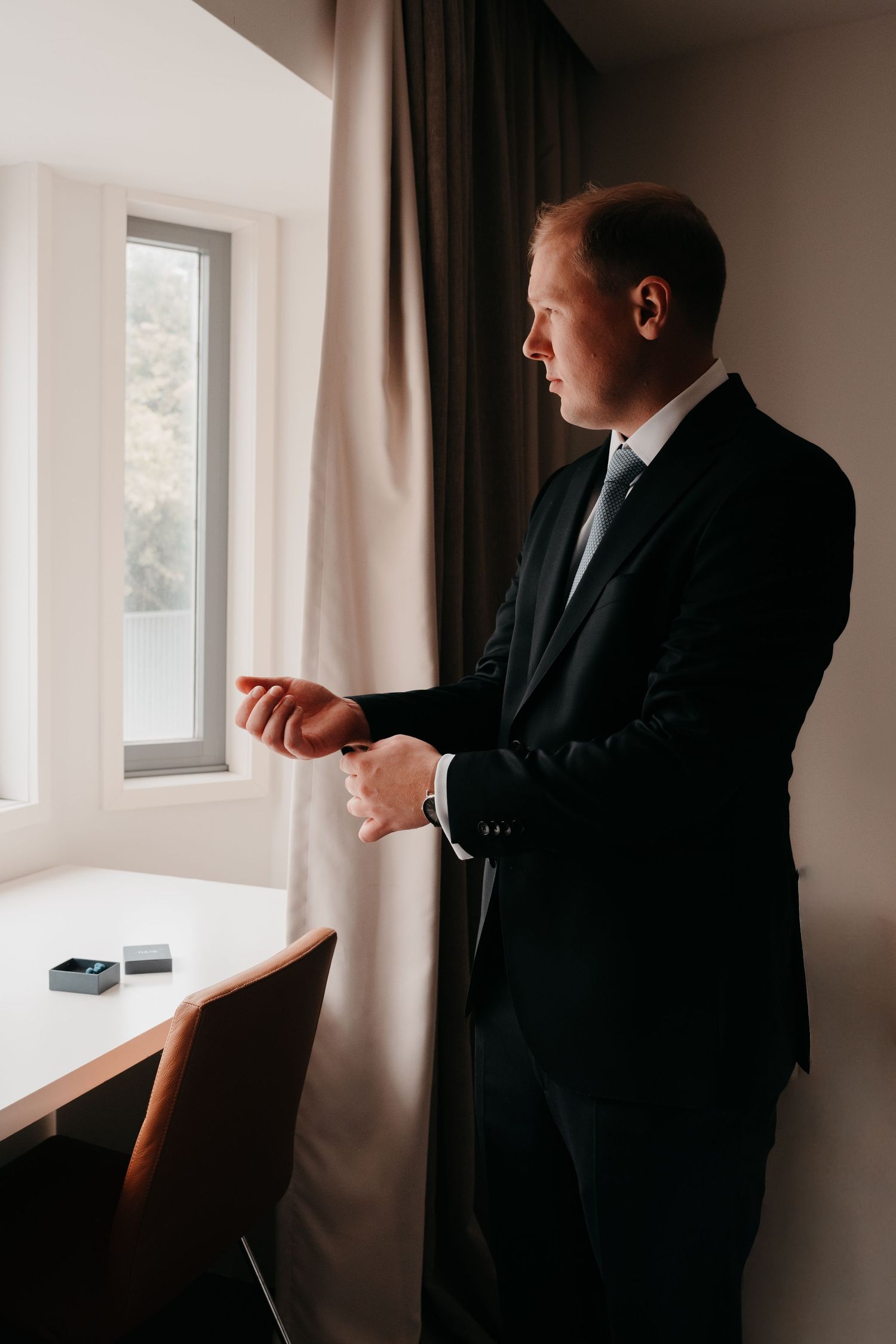 A well-dressed man in a dark suit adjusts his watch cufflinks while standing by a hotel room window.