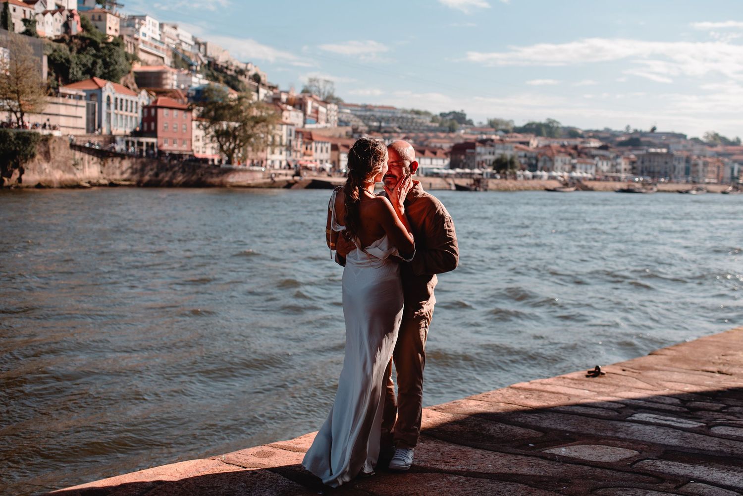 Couple in wedding attire embracing by the Douro River in Porto, Portugal, with colorful buildings in the background.