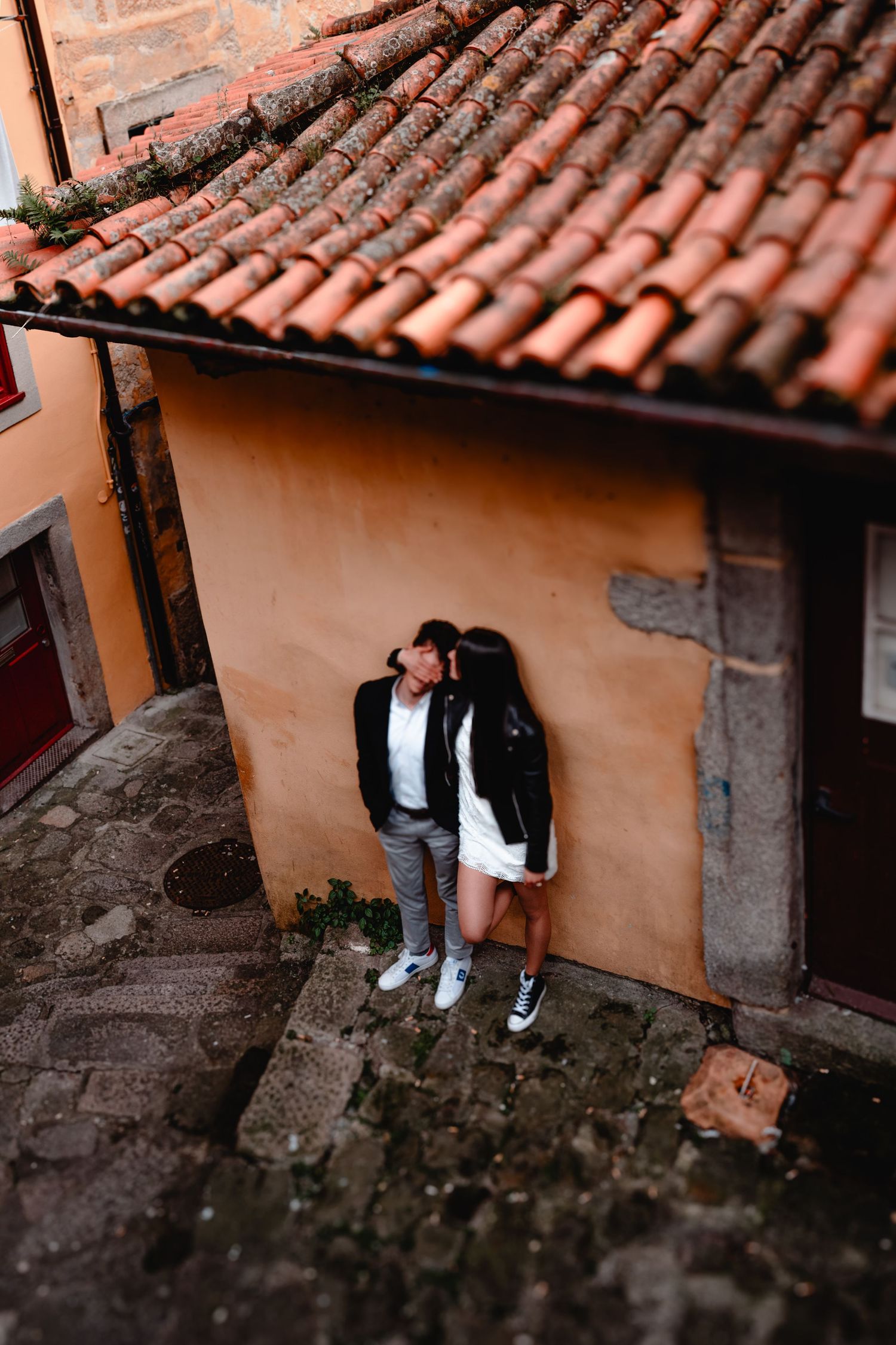 Couple sharing a romantic kiss against an orange wall in a charming European cobblestone alley with terracotta roof tiles.