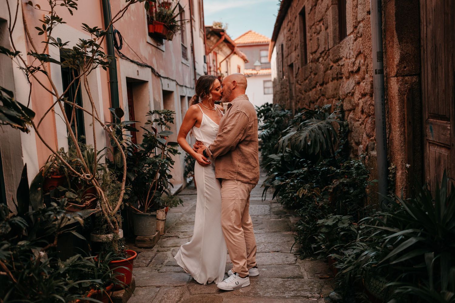 Bride and groom share a romantic embrace in a charming narrow alley lined with lush green plants on their wedding day.