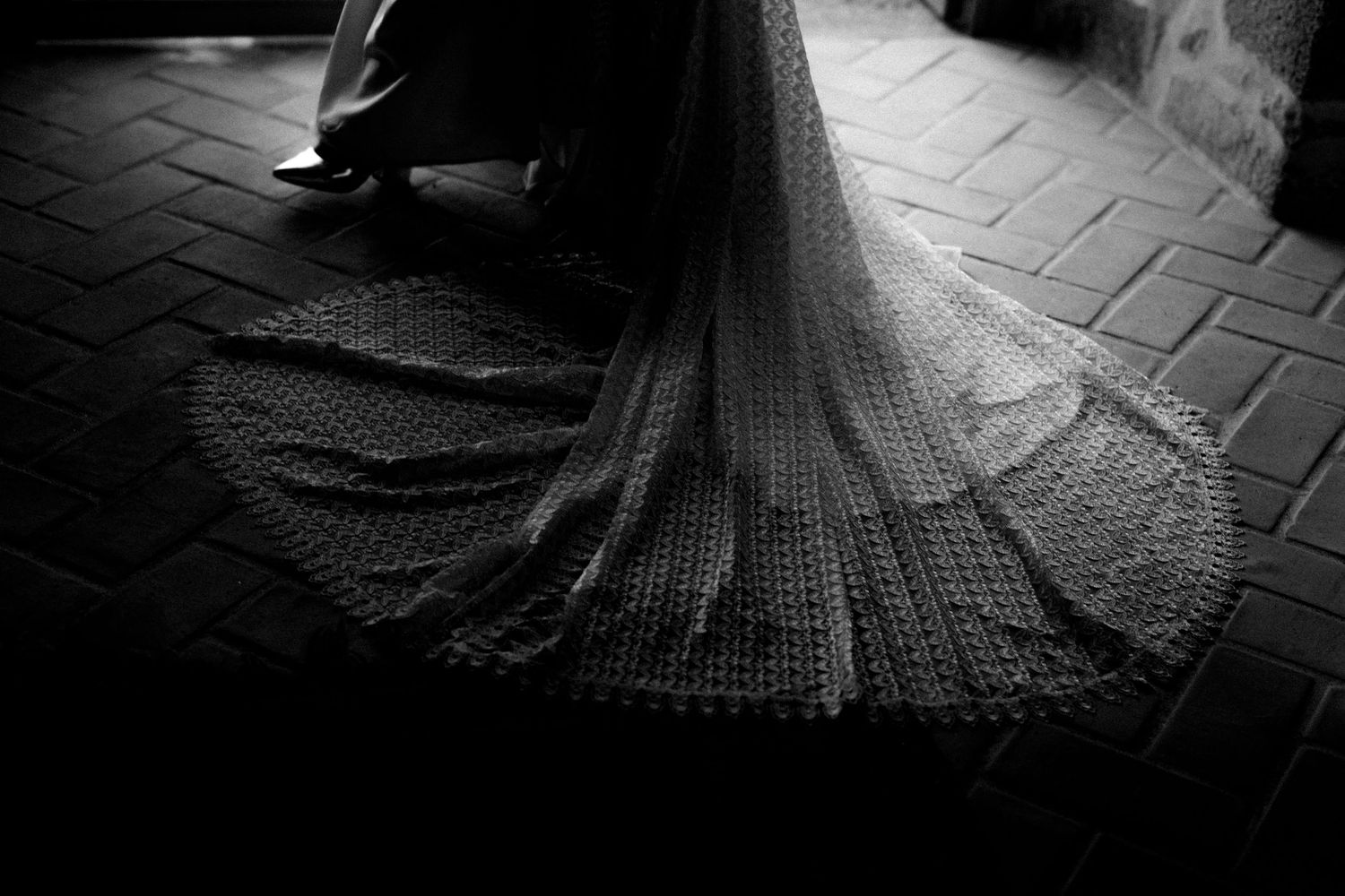 Black and white photo of a bride's lace wedding dress train spread on brick floor.