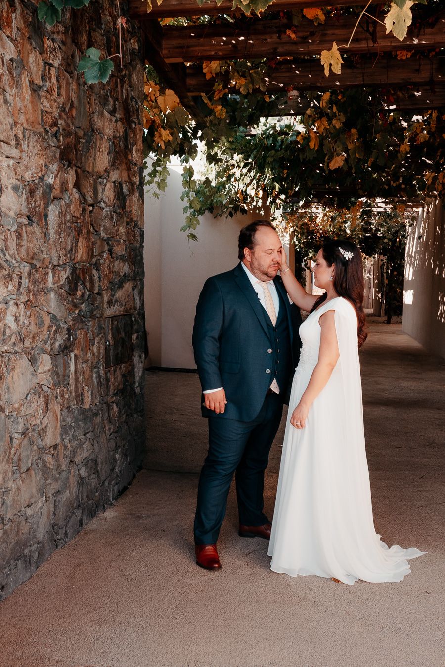 Bride in white gown and groom in navy suit share an intimate moment under a vine-covered pergola beside a stone wall.