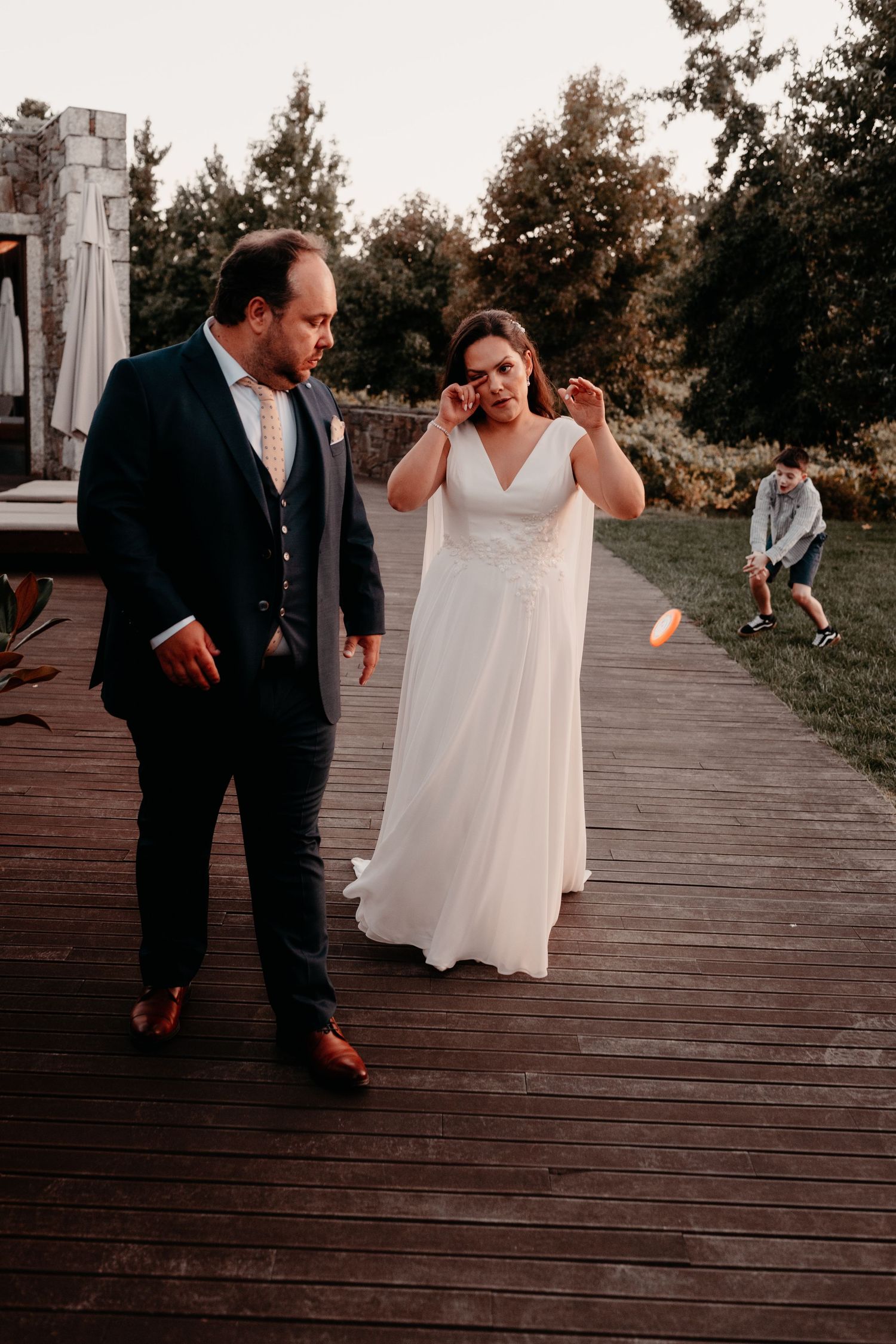Bride and groom walking on wooden deck at sunset, bride wiping tears, child playing in background.