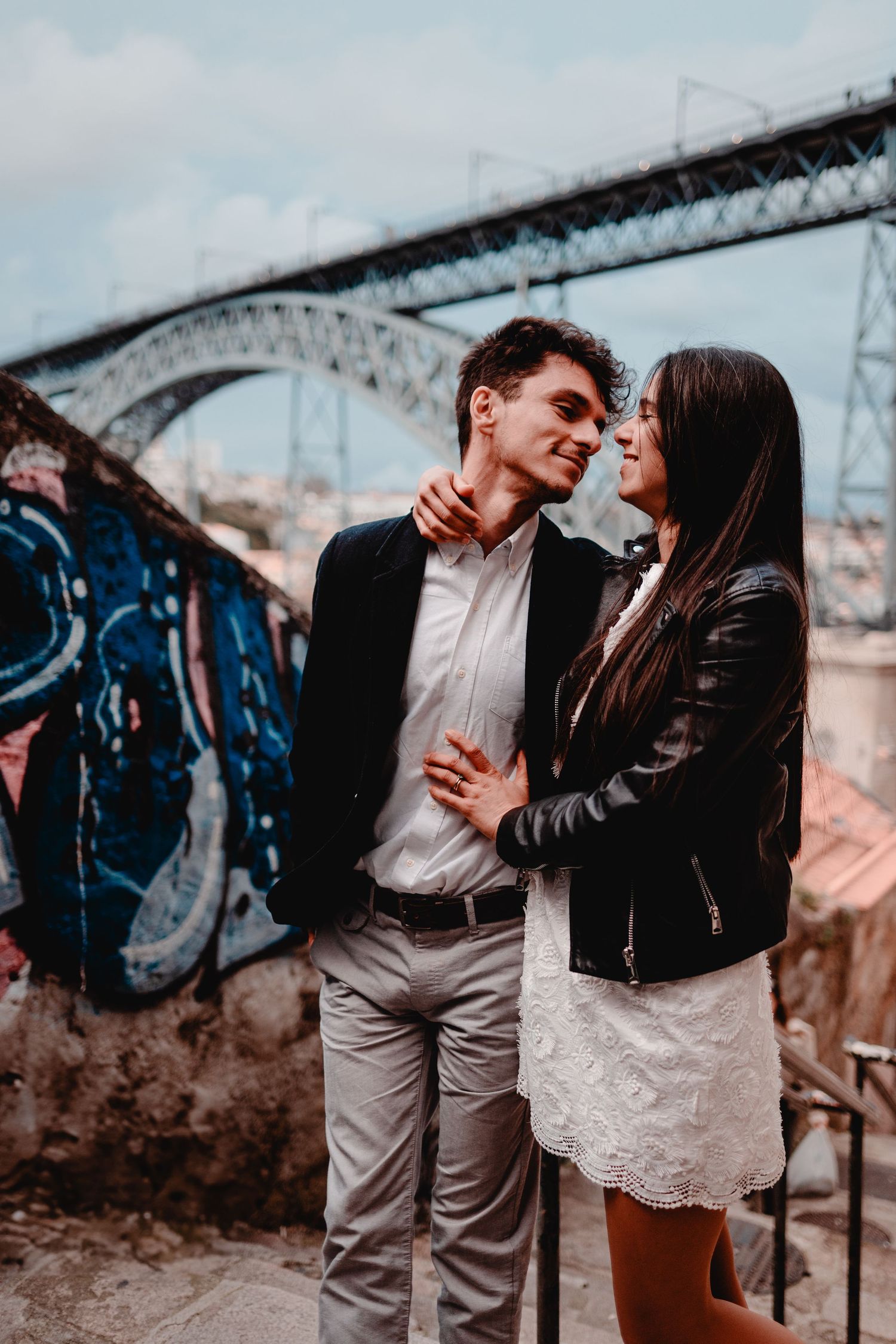 Romantic couple embracing near graffiti wall with iron bridge backdrop in Porto, Portugal.