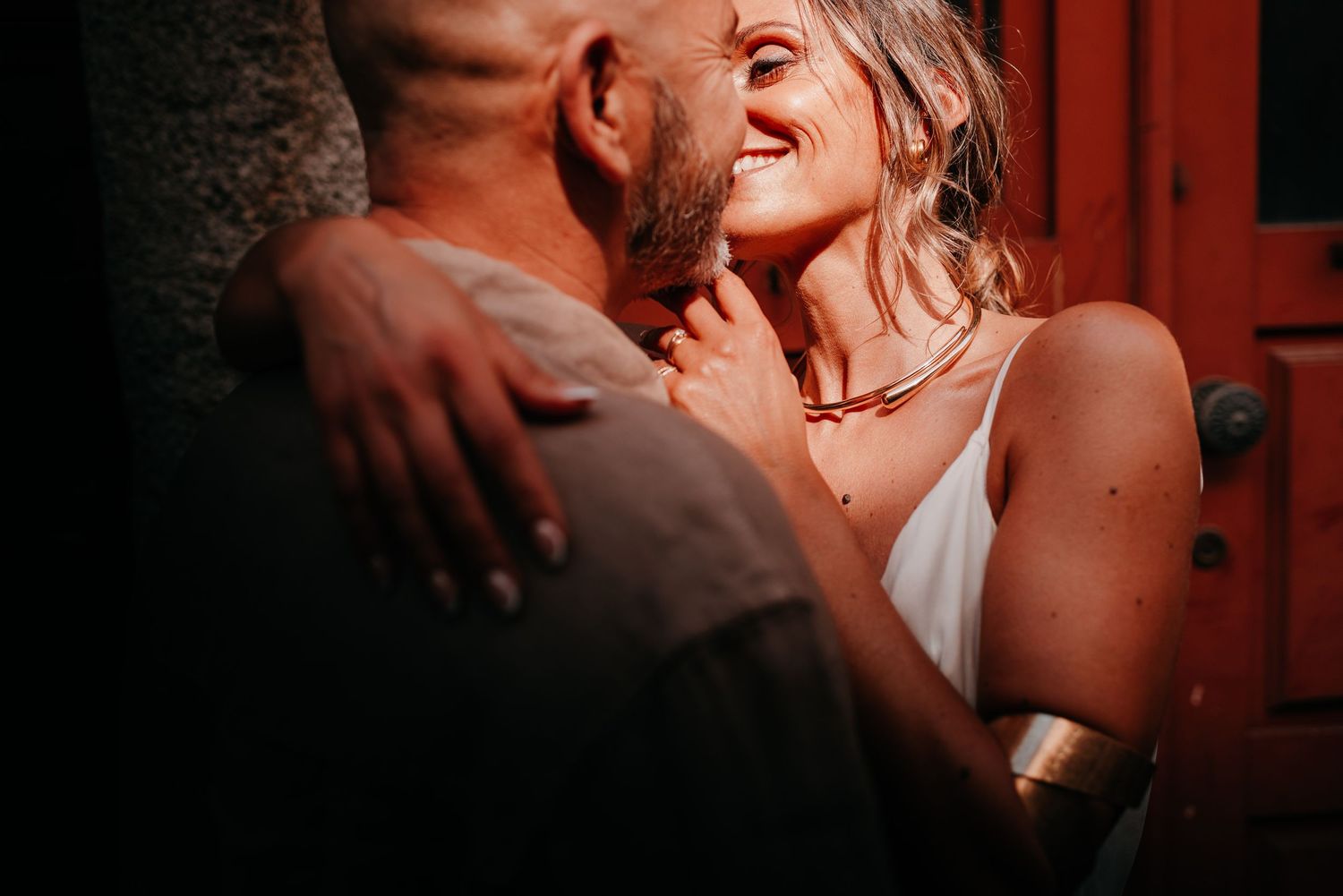 A couple sharing an intimate moment, woman smiling warmly wearing a gold necklace and white dress.