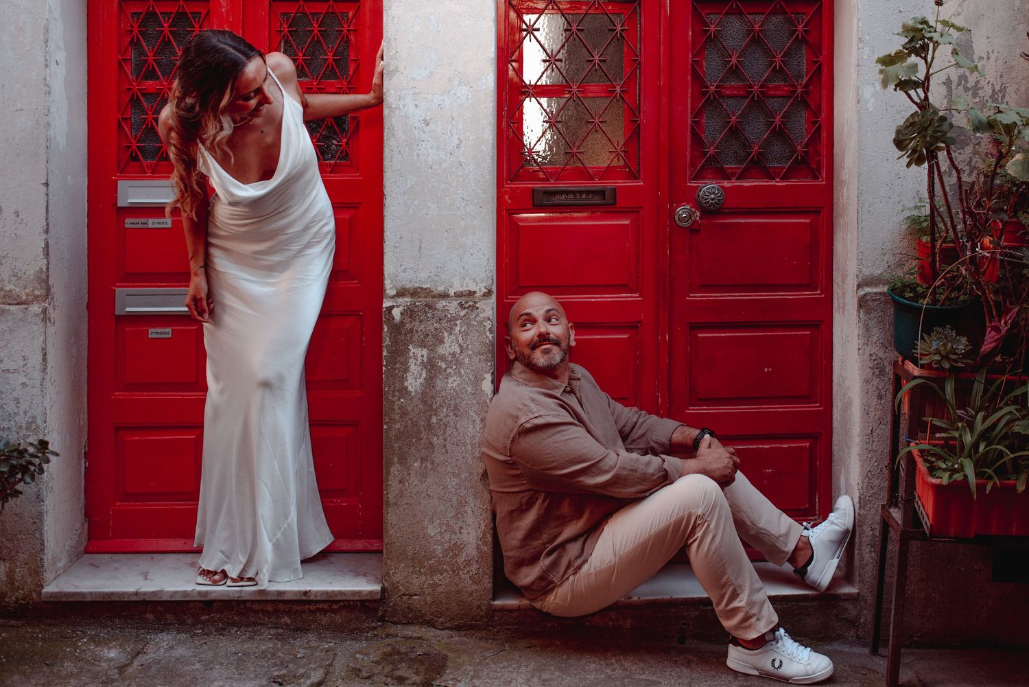 Couple posing playfully against a wall with vibrant red doors and white stone architecture.