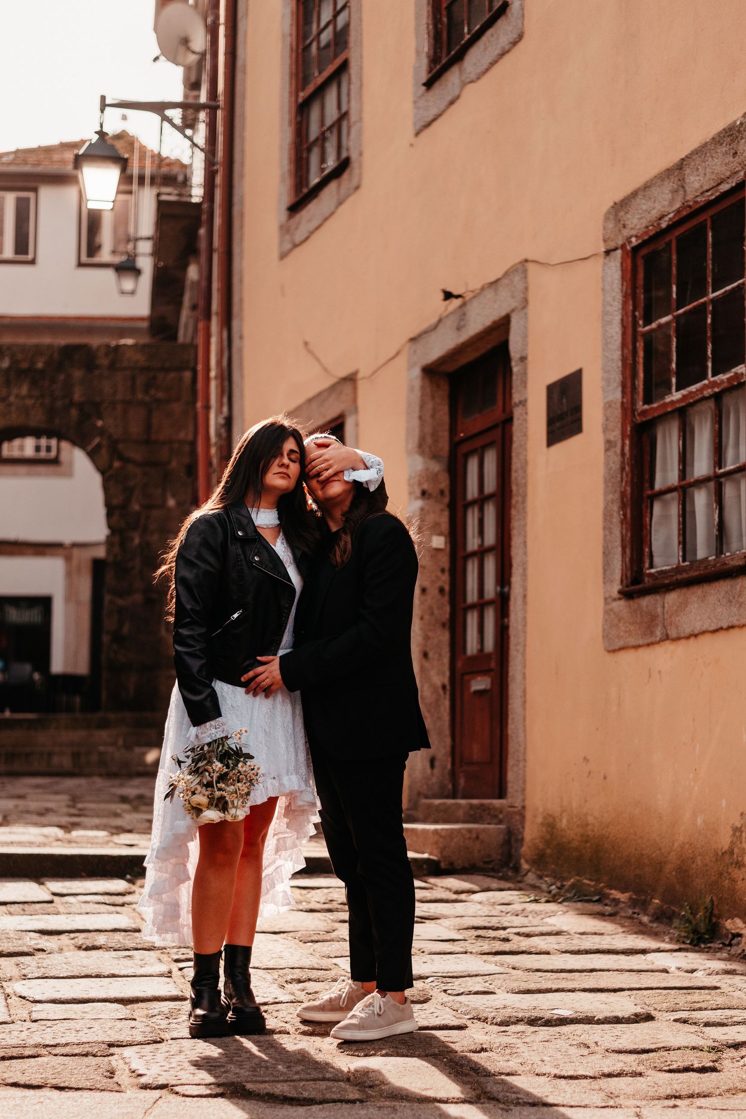 Couple in wedding attire posing on cobblestone street in historic town, bride holding bouquet in white dress and leather jacket.