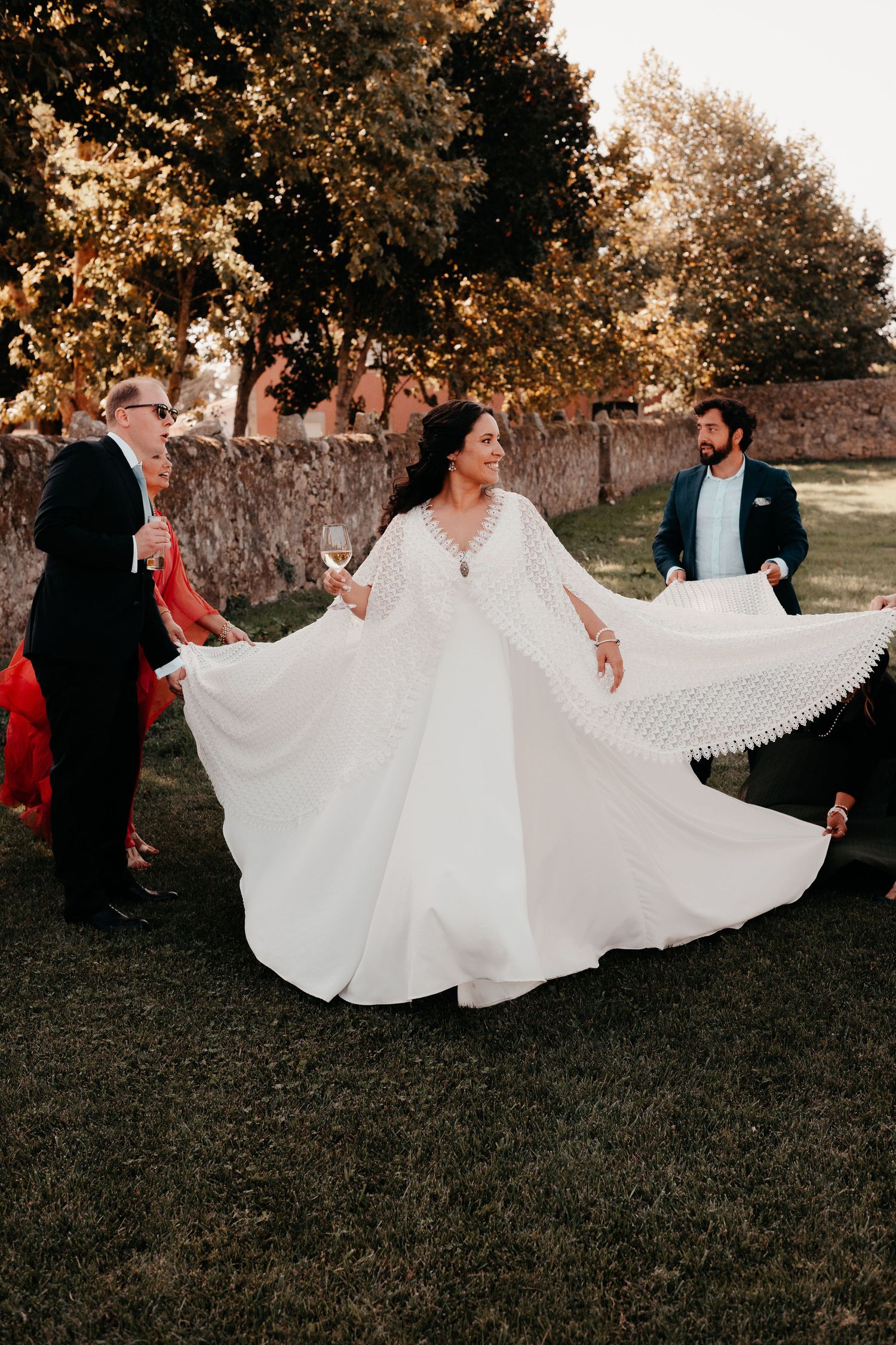 Bride in flowing white cape wedding dress twirling outdoors with guests on green lawn near stone wall.