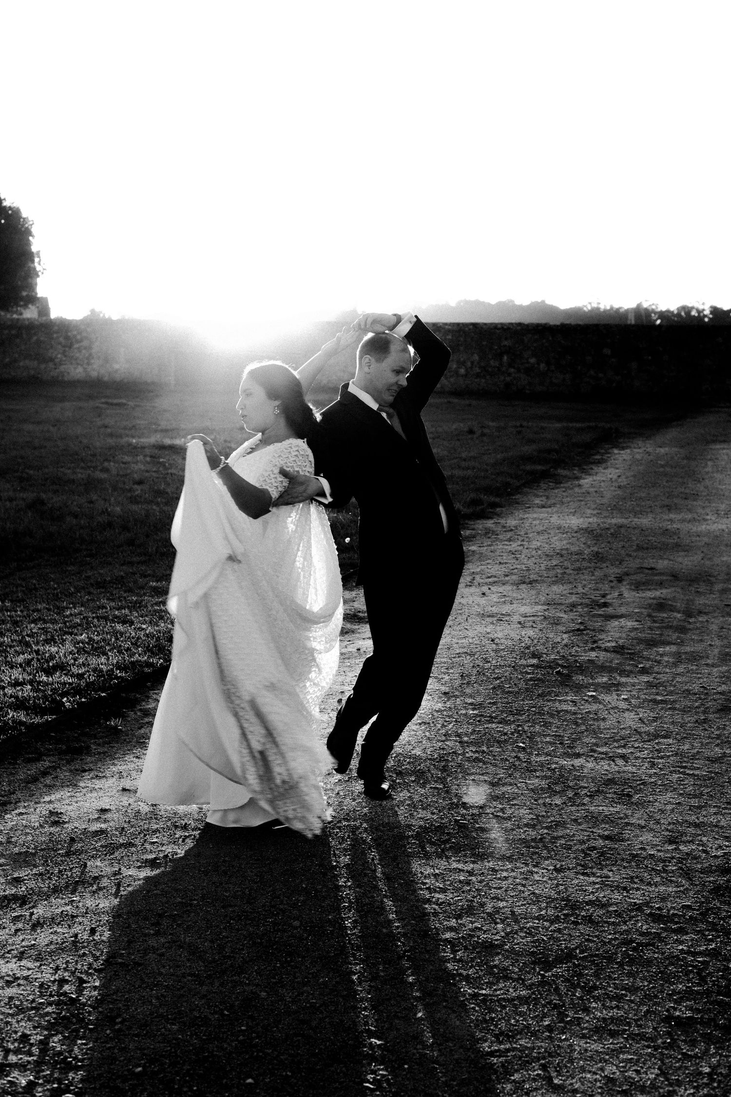 Black and white photo of a bride and groom dancing on a rural path at sunset, casting long shadows.