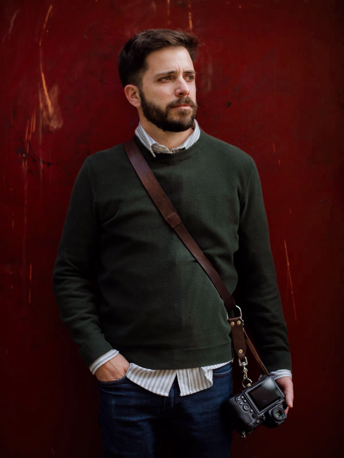 A bearded man in a green sweater carries a camera on a leather strap, posing against a dark red wall.