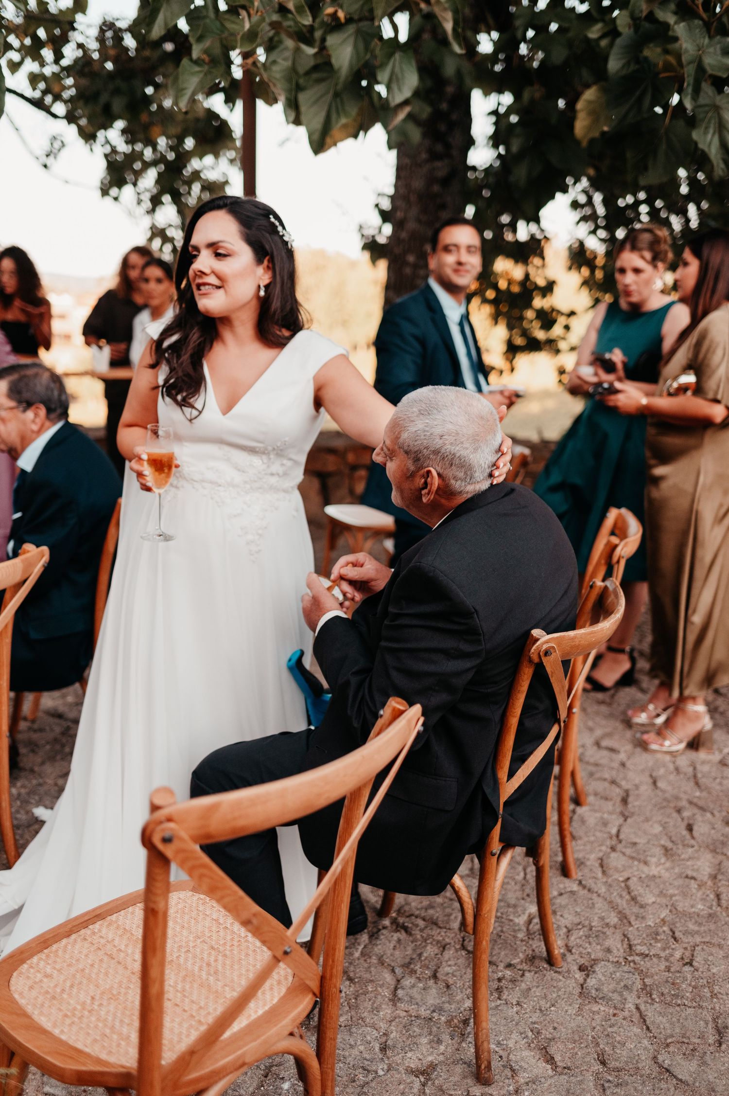 Bride in white gown holding wine glass interacts with seated elderly guest at outdoor wedding reception.
