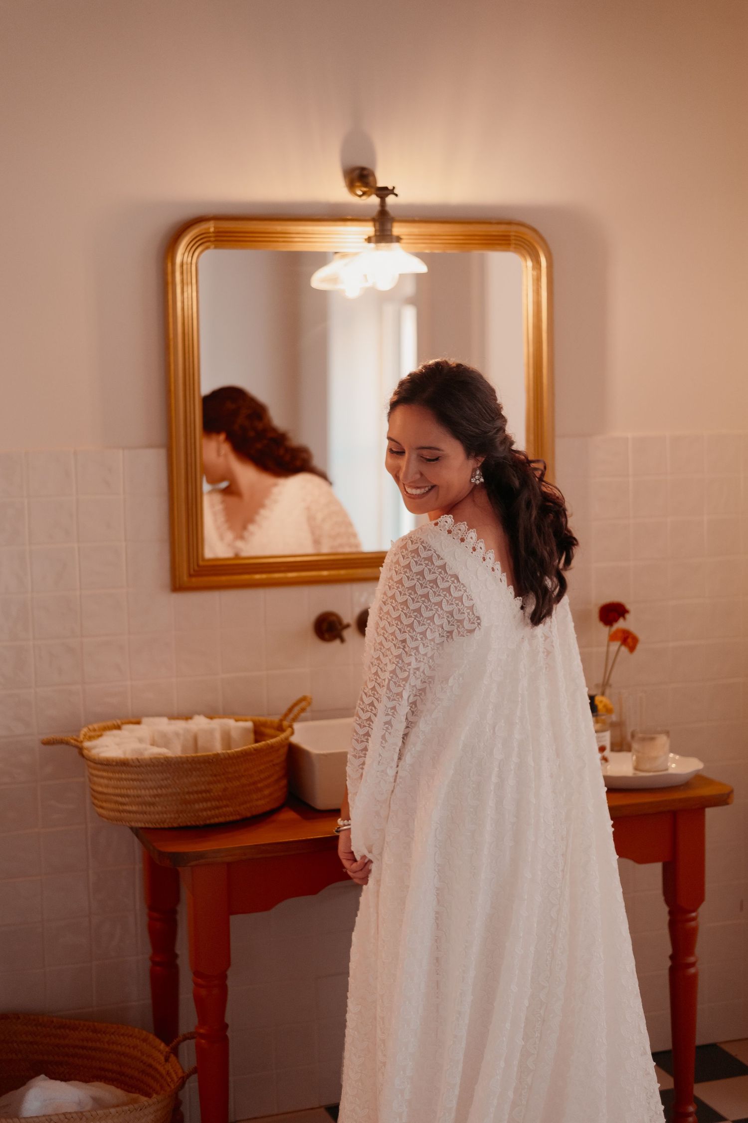 Smiling bride in white lace wedding dress standing in elegant bathroom with gold mirror reflection.