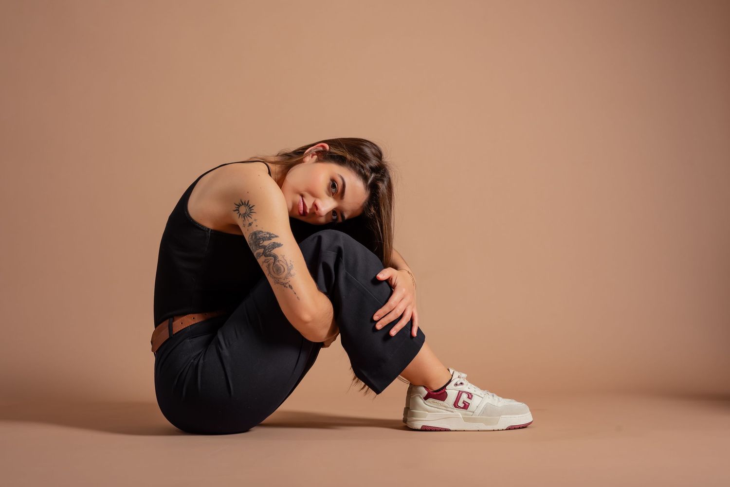 Artistic portrait showing curled up pose against peachy brown backdrop wearing dark casual attire.