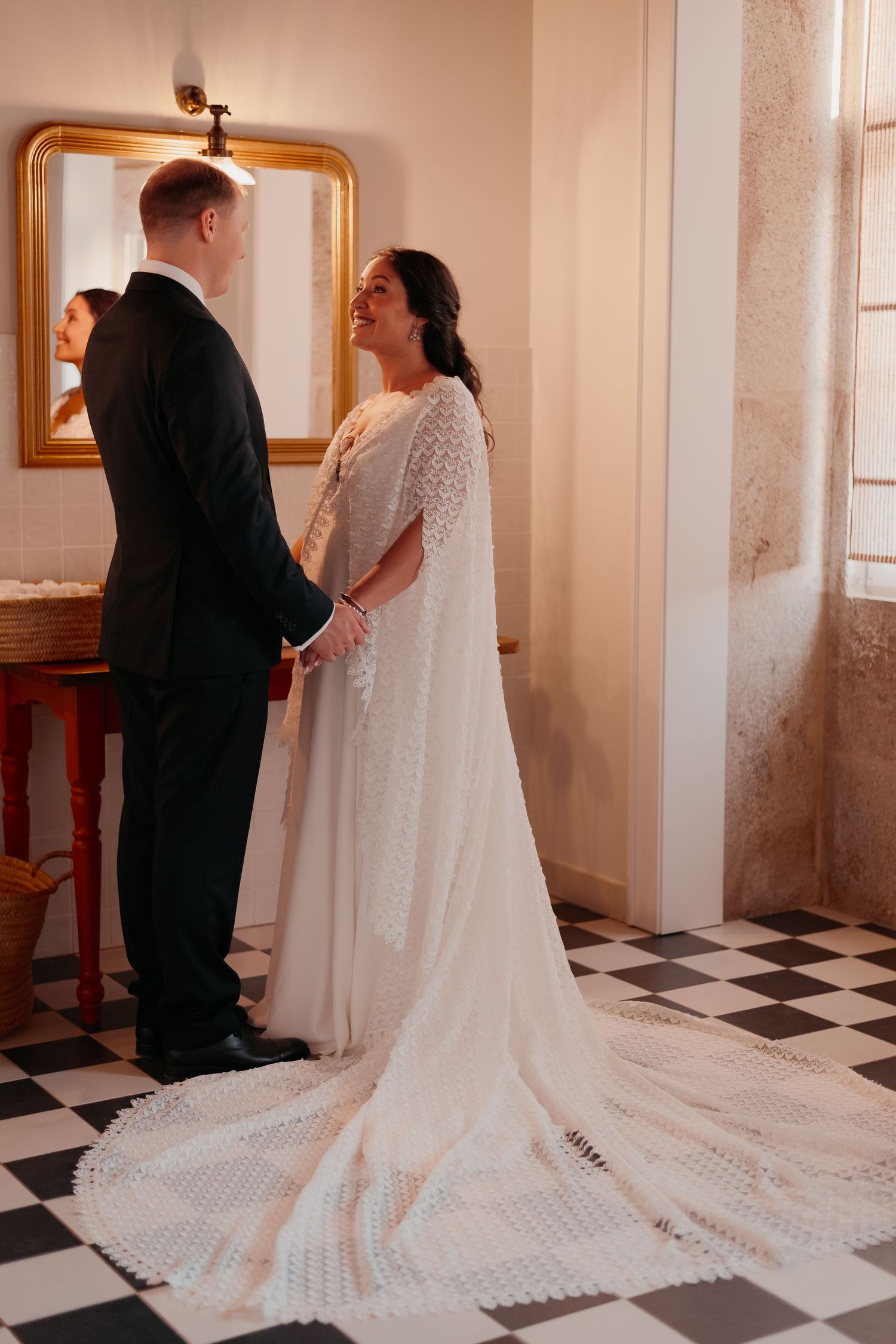 Bride in lace cape wedding gown holds hands with groom in black tuxedo on checkered floor near gold mirror.