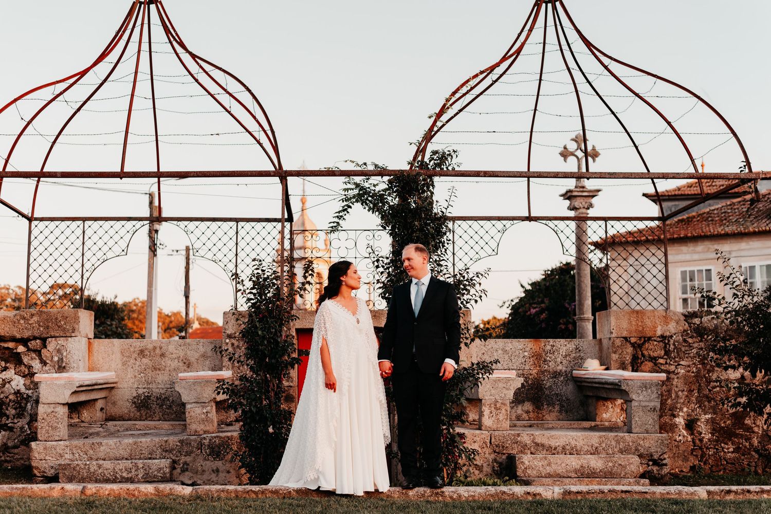 Bride and groom stand together in a romantic garden with ornate metal gazebo structures at golden hour.