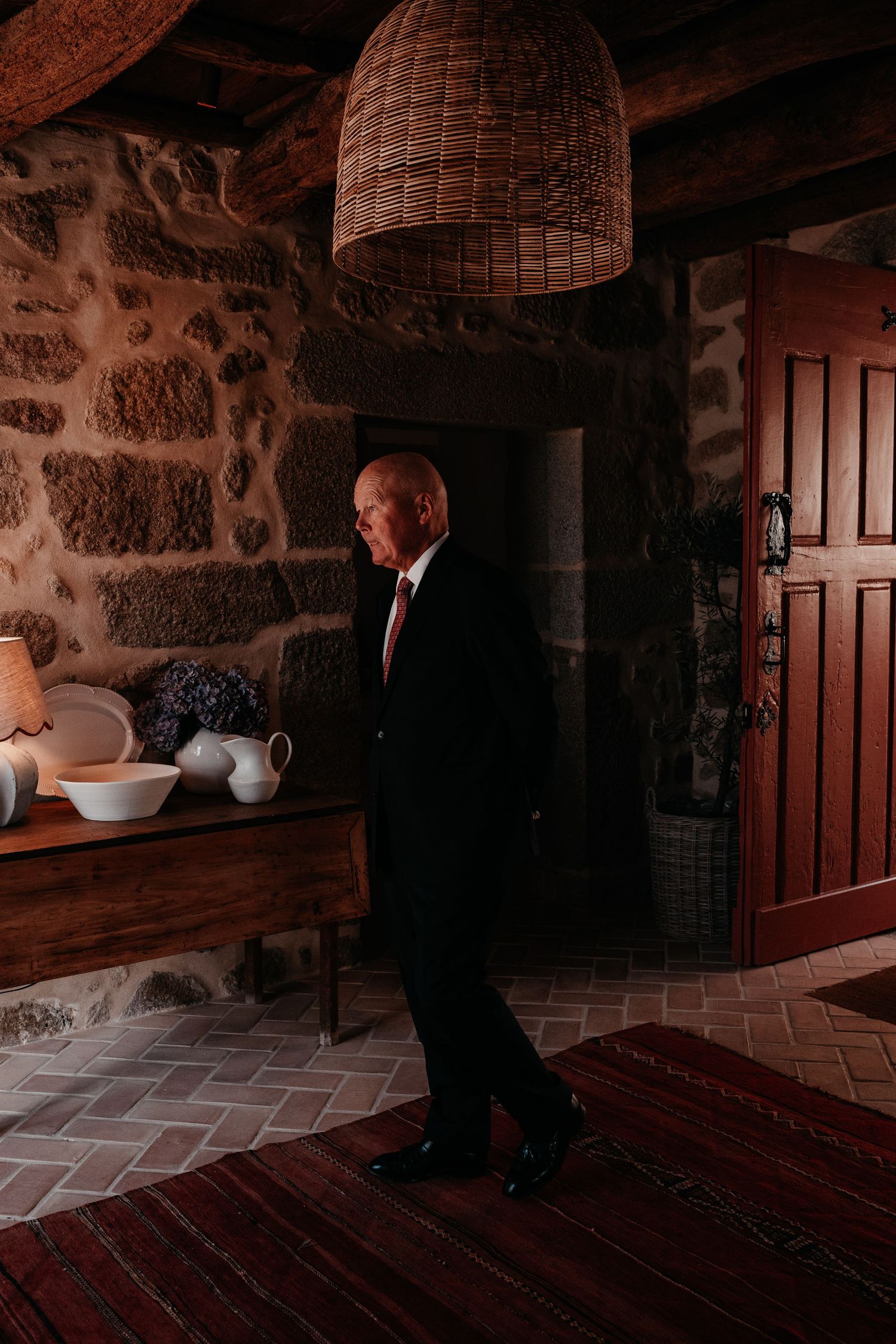 Elderly man in black suit with red tie standing in rustic stone interior with wooden furniture and wicker pendant light.