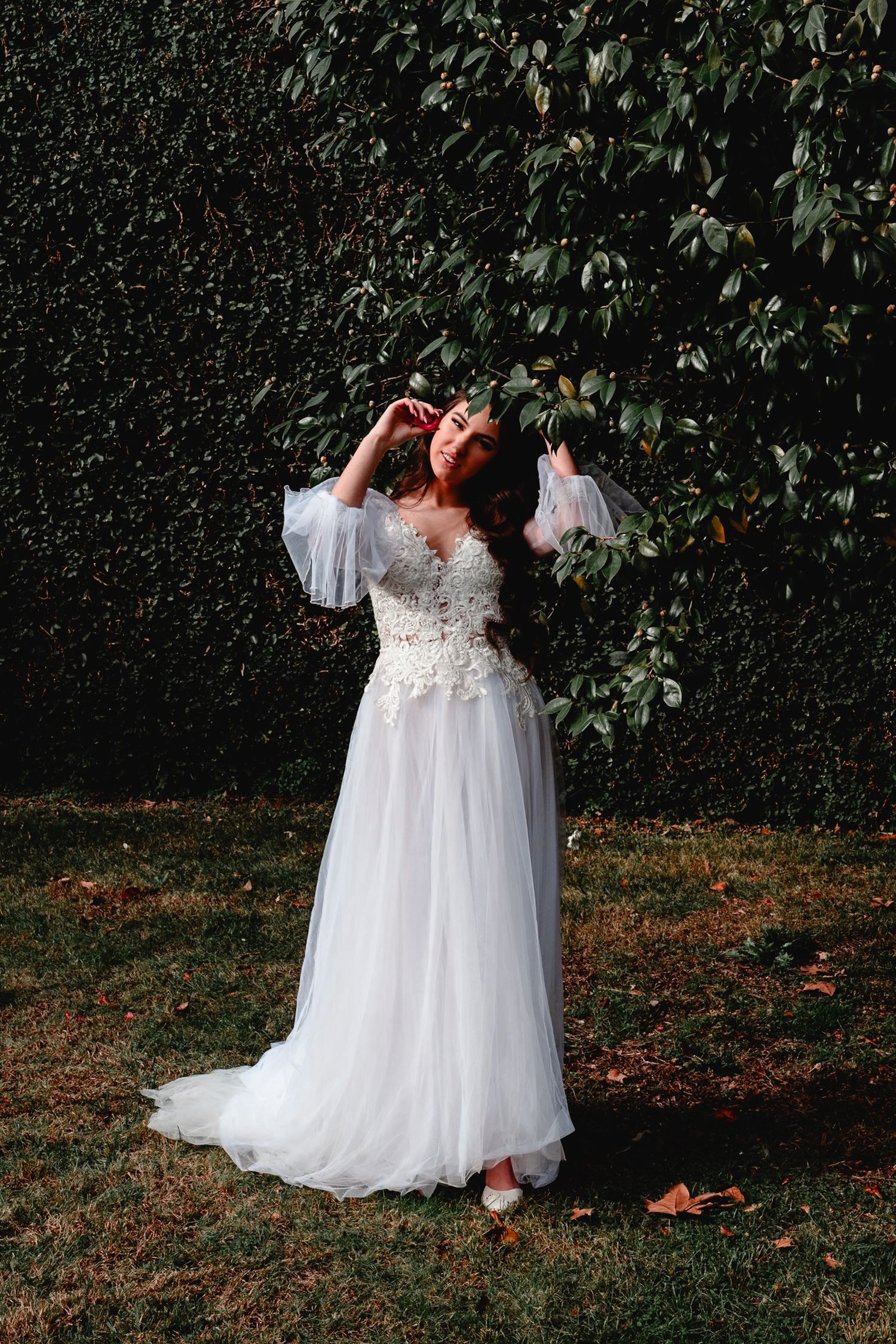 A bride in a flowing white lace gown poses gracefully against a lush green hedge backdrop in a garden setting.