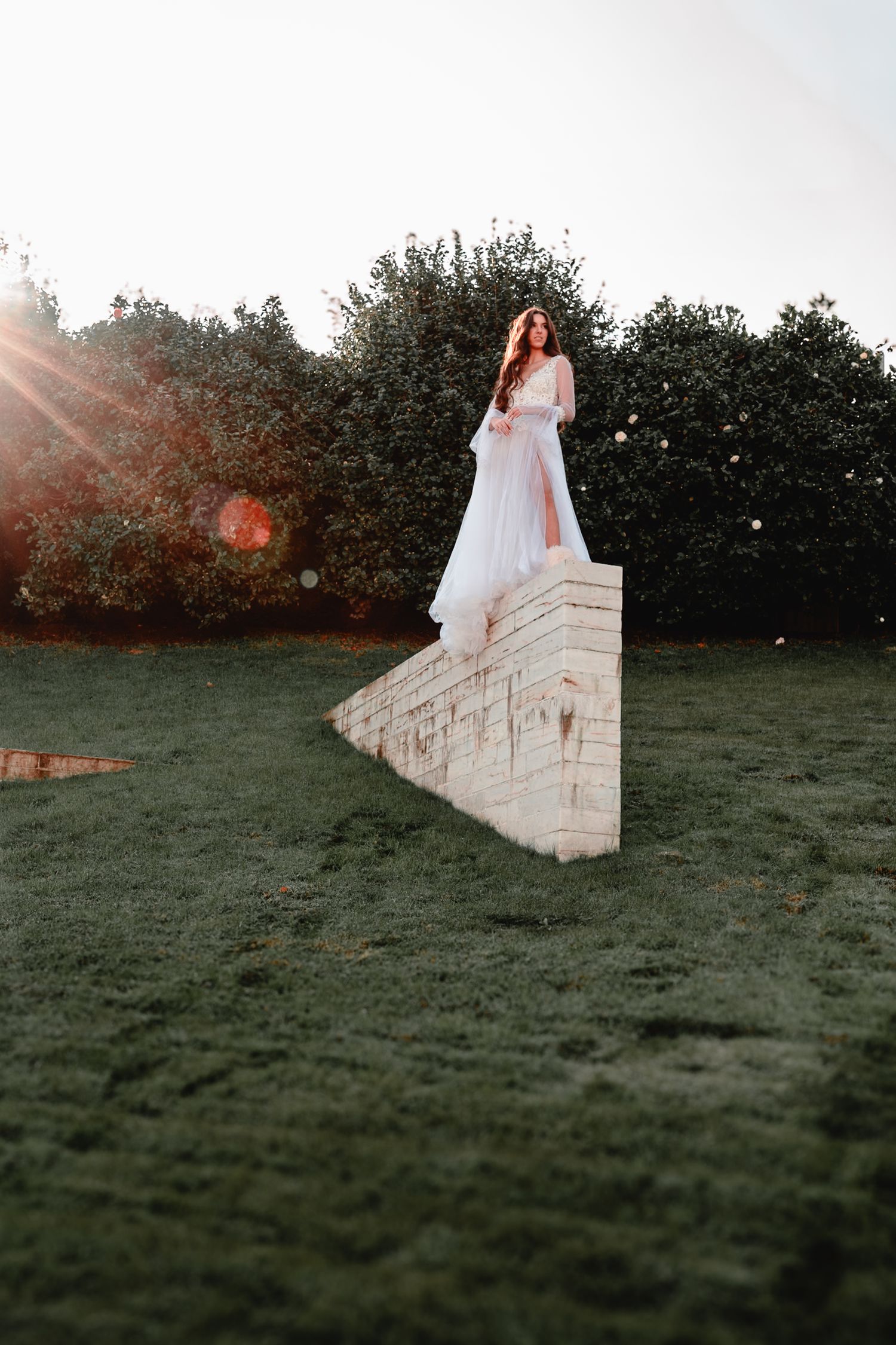 Dramatic bridal portrait on stone wall with sunflare and garden backdrop at golden hour.