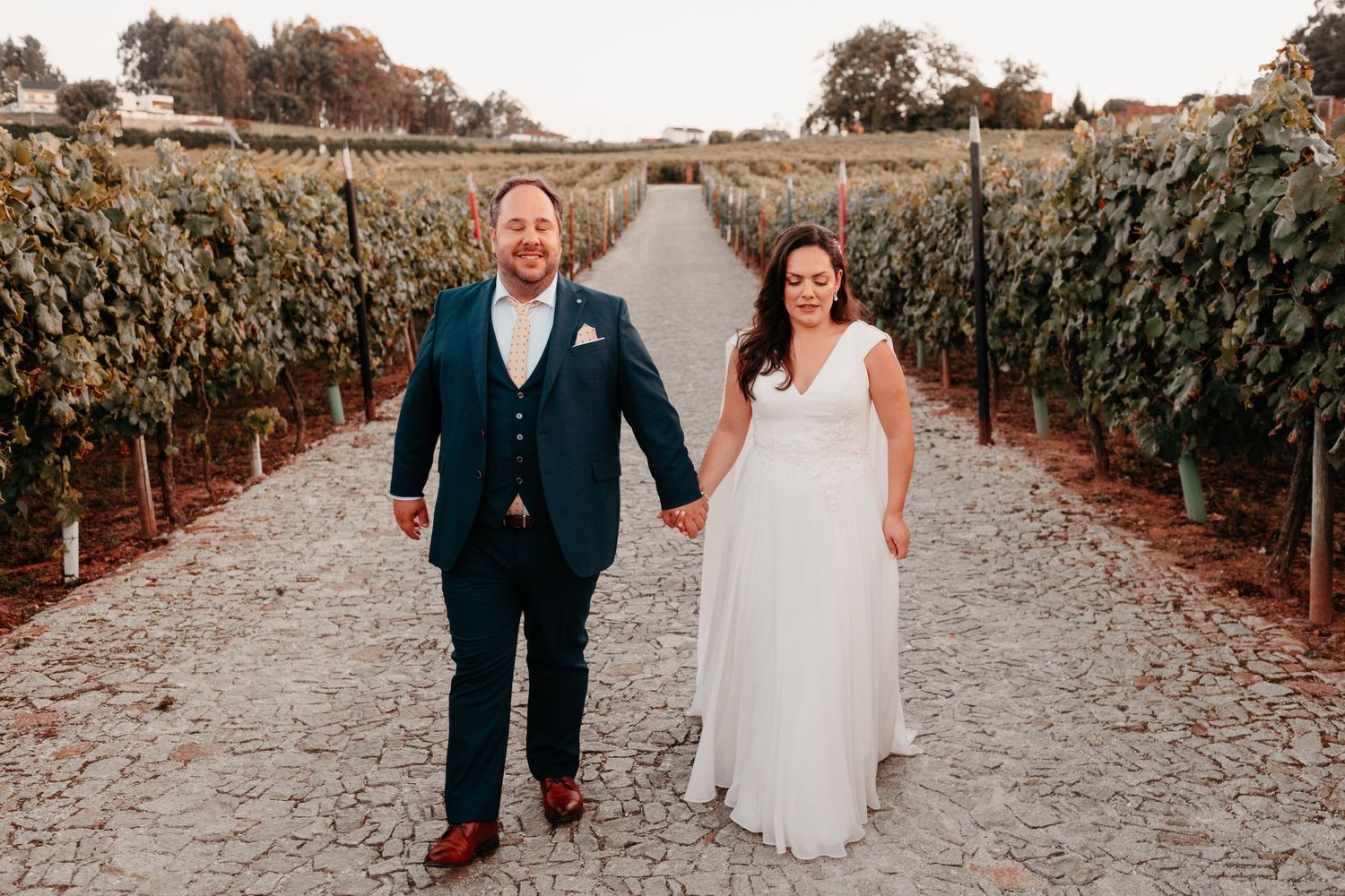 Bride and groom holding hands walking through vineyard rows, bride in white gown, groom in teal suit.